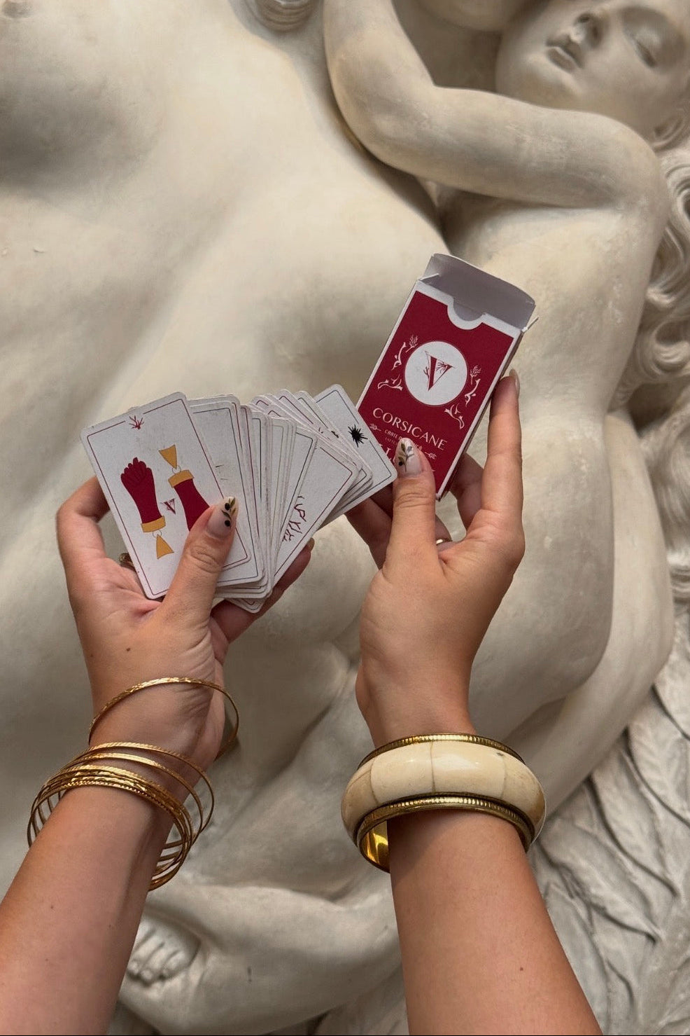A person's hands holding a deck of mediterranean cards and fanning them out in front of a large white marble sculpture. The cards and box are red and white with gold accents.