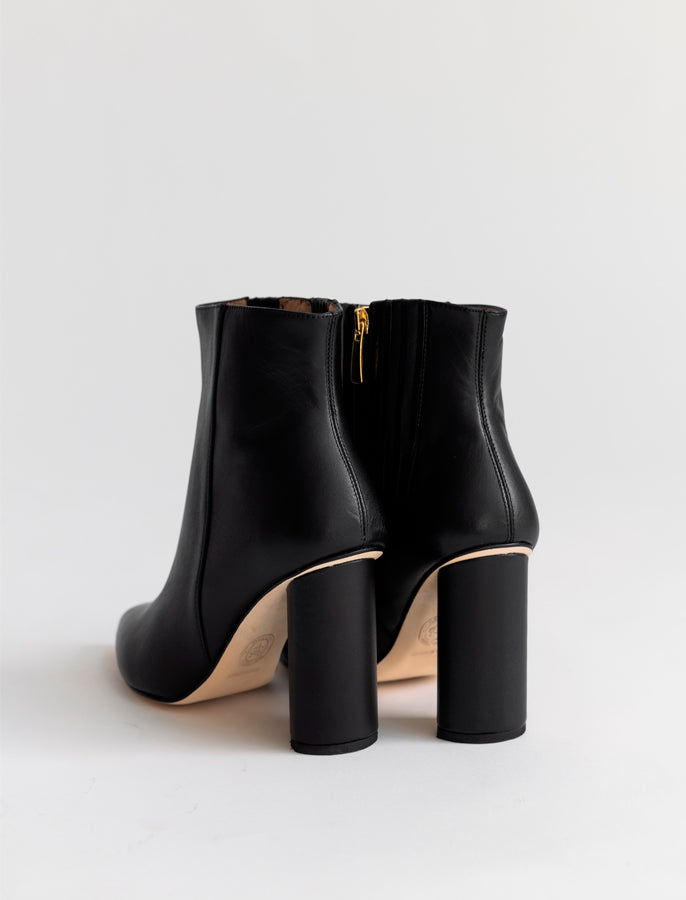 A pair of black leather ankle boots with chunky block heels and a gold zipper, shown from a rear three-quarter angle on a light gray background.