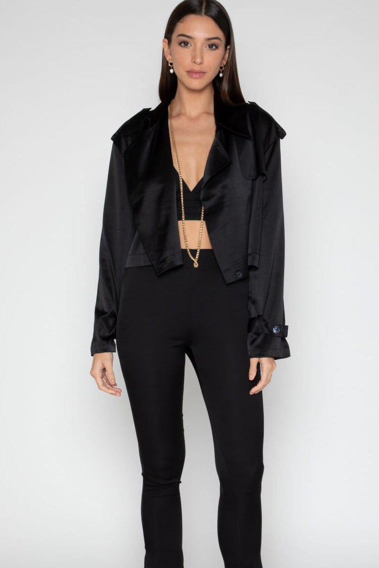 A woman modeling a black satin cropped jacket with a wide collar and black high-waisted pants against a light gray studio background.