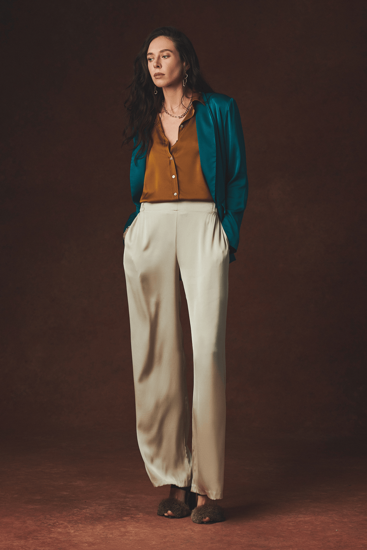 A woman stands against a dark brown studio backdrop wearing a teal open-front jacket over a rust-colored blouse, paired with wide-leg cream trousers and dark brown shoes.