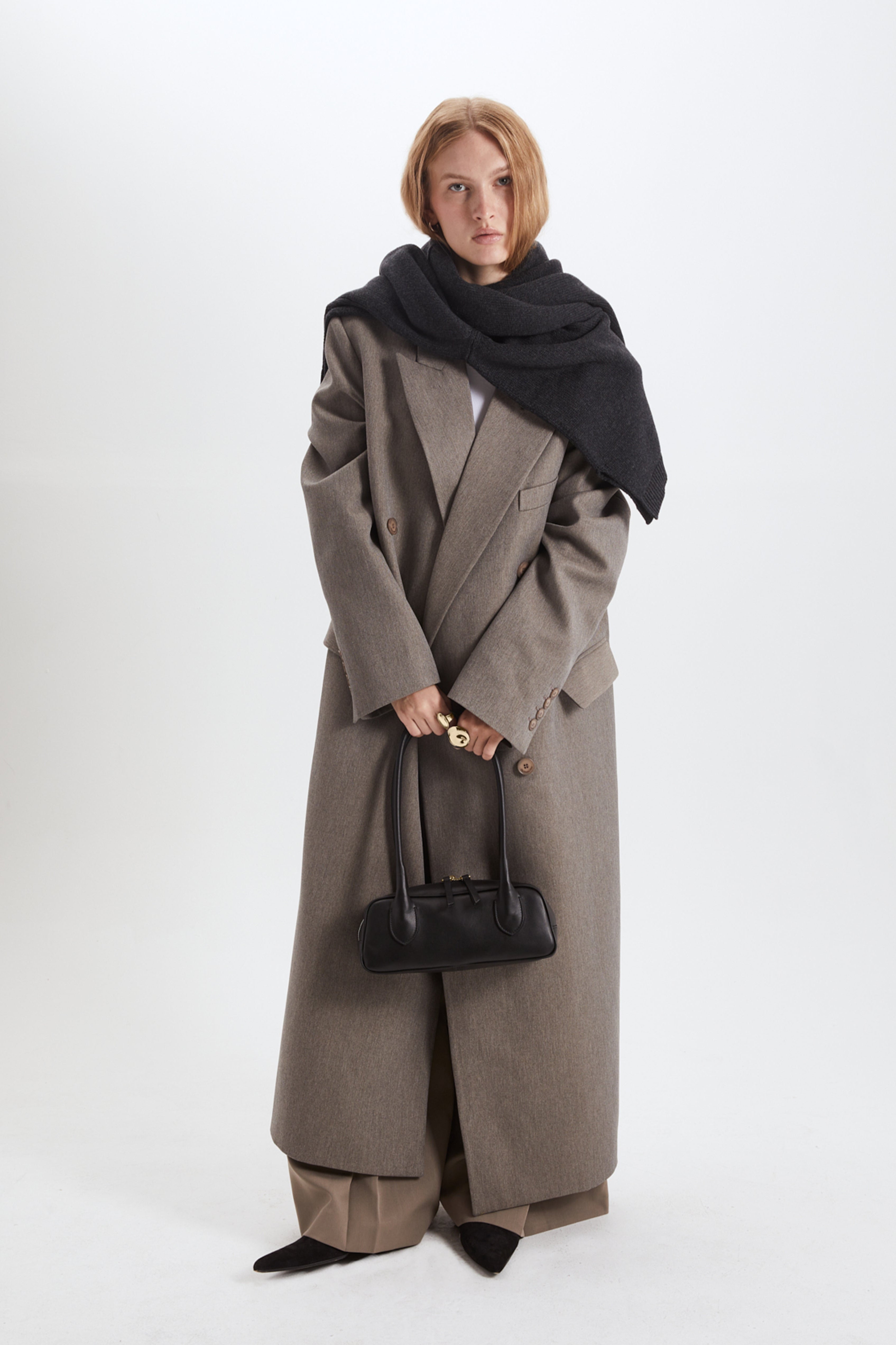Person wearing a long gray coat and black scarf holding a black handbag on a white background
