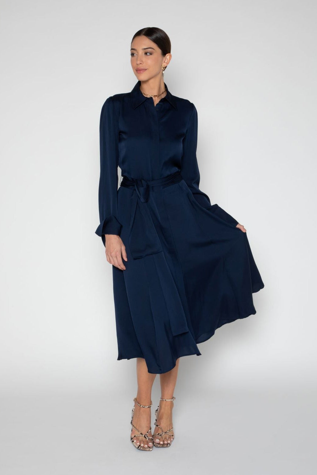 A woman modeling a dark navy blue long-sleeve midi dress with a shawl collar, tie waist, and flared skirt, paired with metallic high-heeled sandals against a light gray studio background.