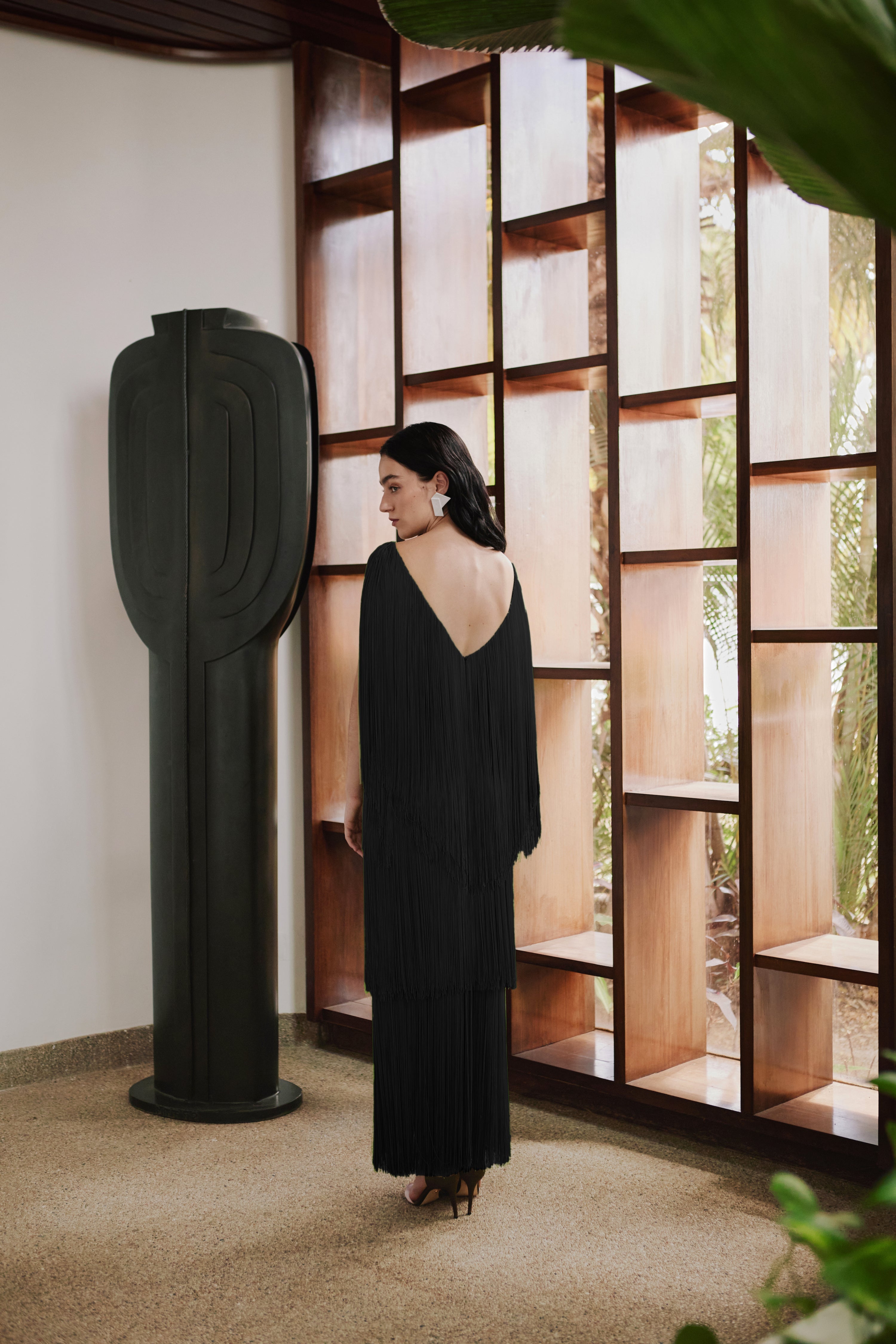 Woman in an open-back black dress standing in a room with a modern sculpture and large windows.