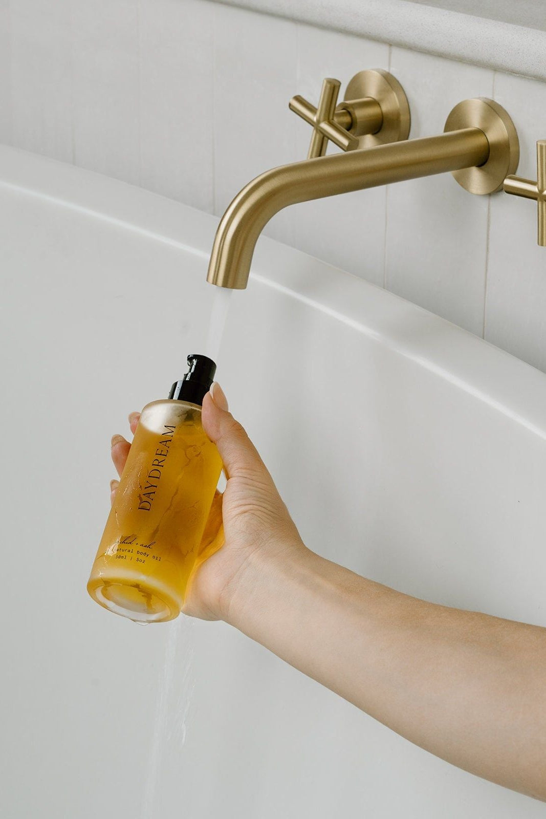 Hand holding a bottle of liquid next to a gold faucet in a bathroom setting