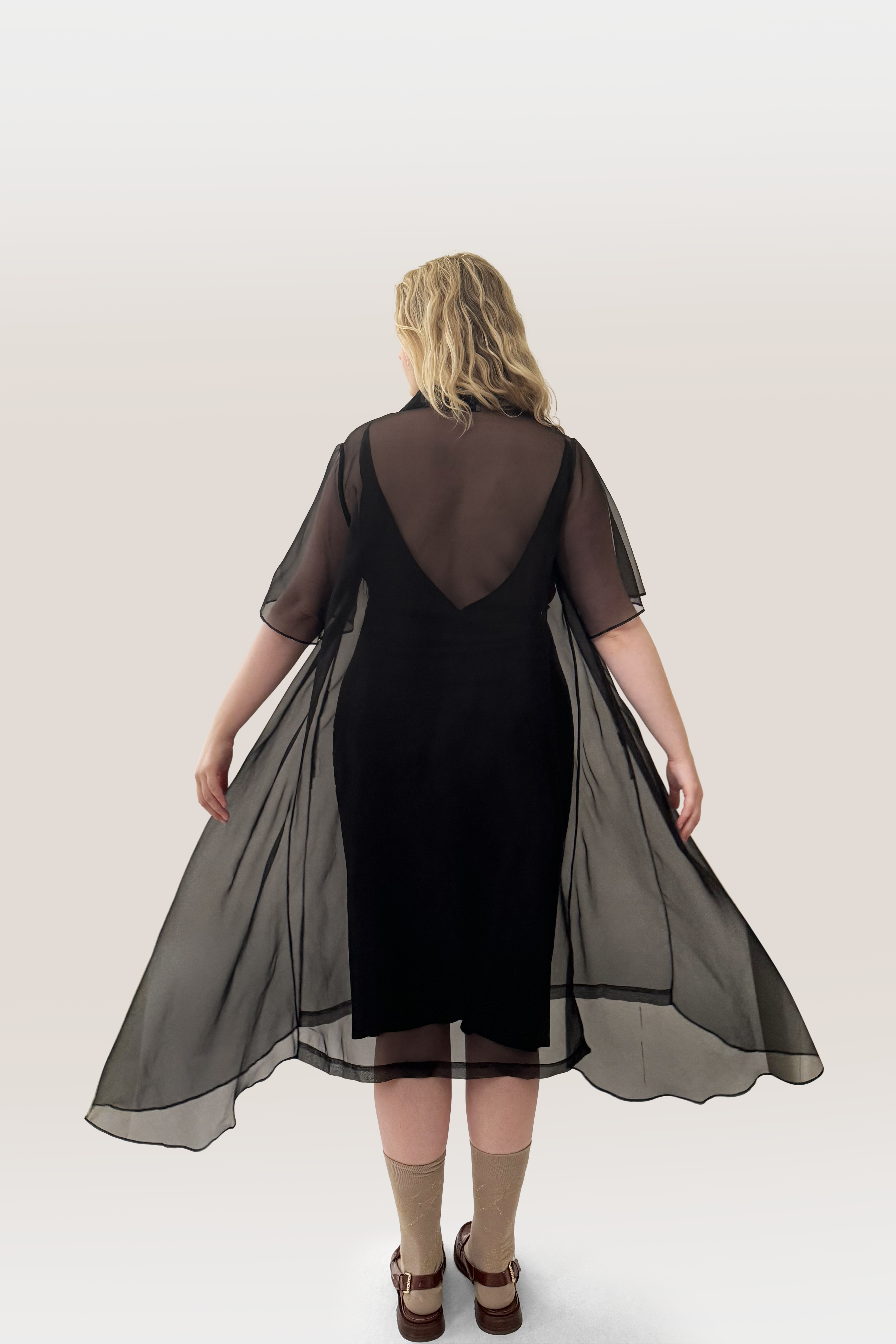 A woman viewed from behind wearing a black midi silk slip dress with a V-shaped back and a sheer black silk overlay cape featuring short sleeves, set against a plain light gray studio background.