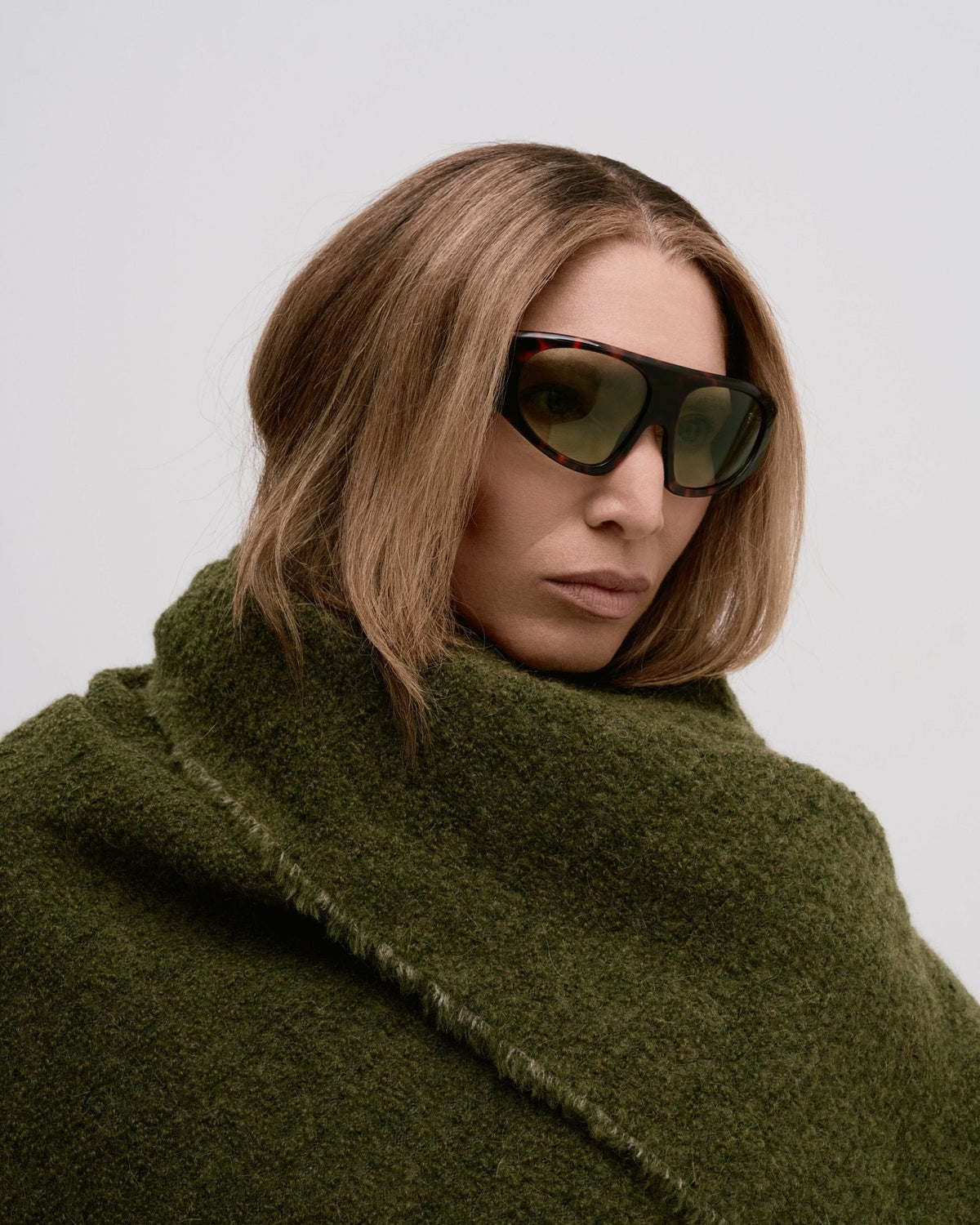 Woman wearing sunglasses and an olive green throw blanket wrapped around her against a light gray background