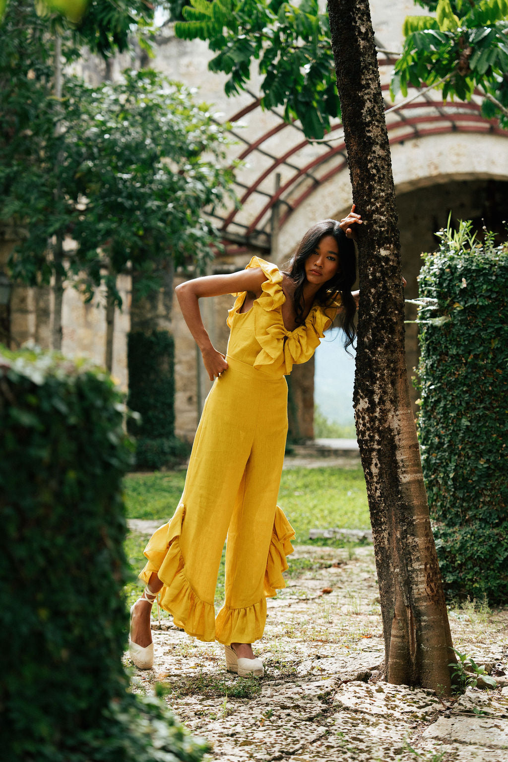 Woman in a yellow dress standing in a garden with trees and arches in the background