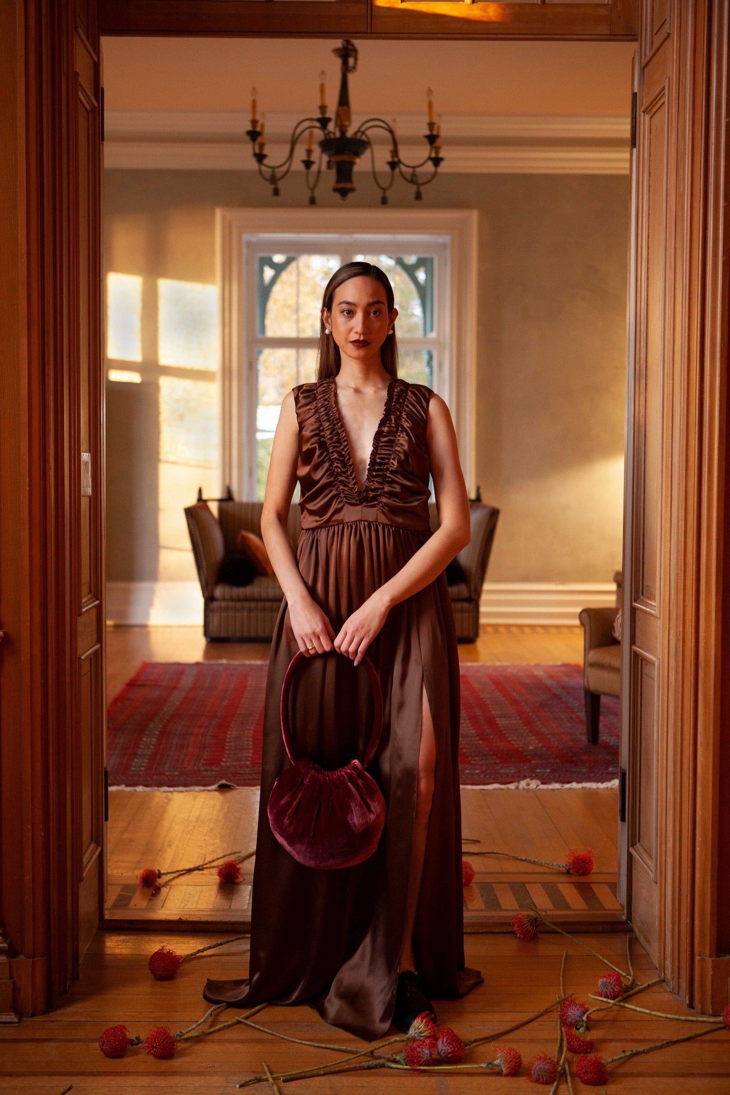 Woman in a long brown silk dress holding a matching velvet handbag in a room with a chandelier and large window.