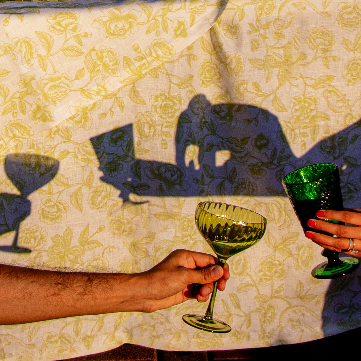 Two people holding green glasses with a floral tablecloth in the background