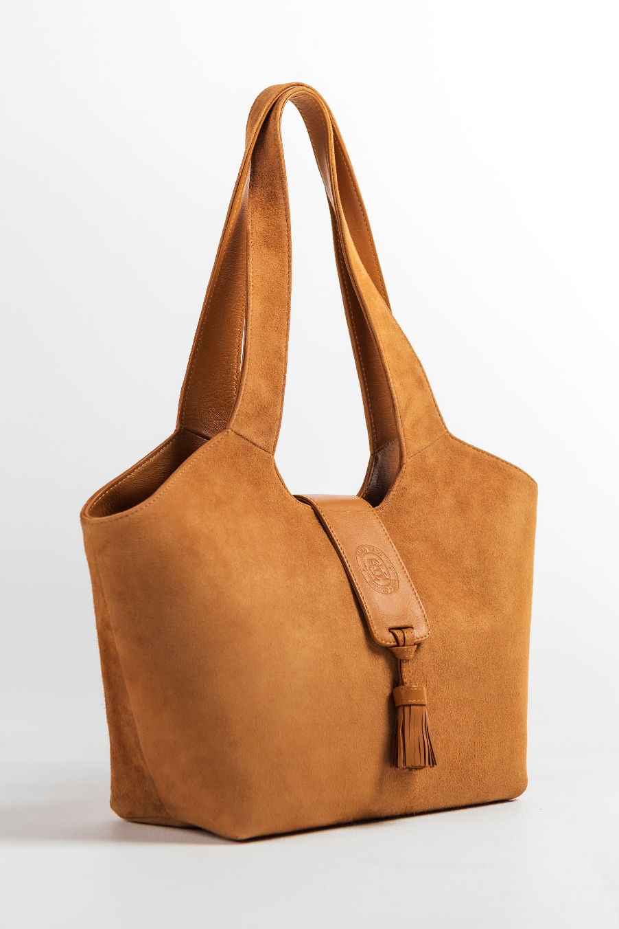 A tan suede tote bag with dual shoulder straps, a front strap detail, and a tassel accent, photographed on a clean white background.