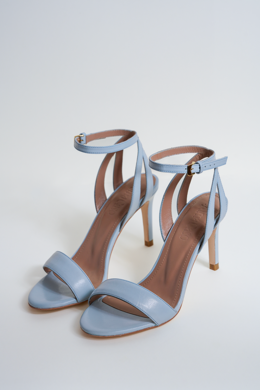 A pair of light blue leather high-heeled sandals with thin ankle straps and open toes, displayed on a clean light gray studio background.