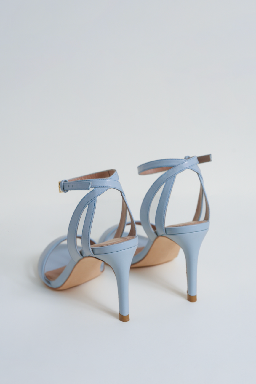 A pair of light blue leather stiletto sandals with thin wrap-around ankle straps, shown from a rear three-quarter angle on a clean white background.