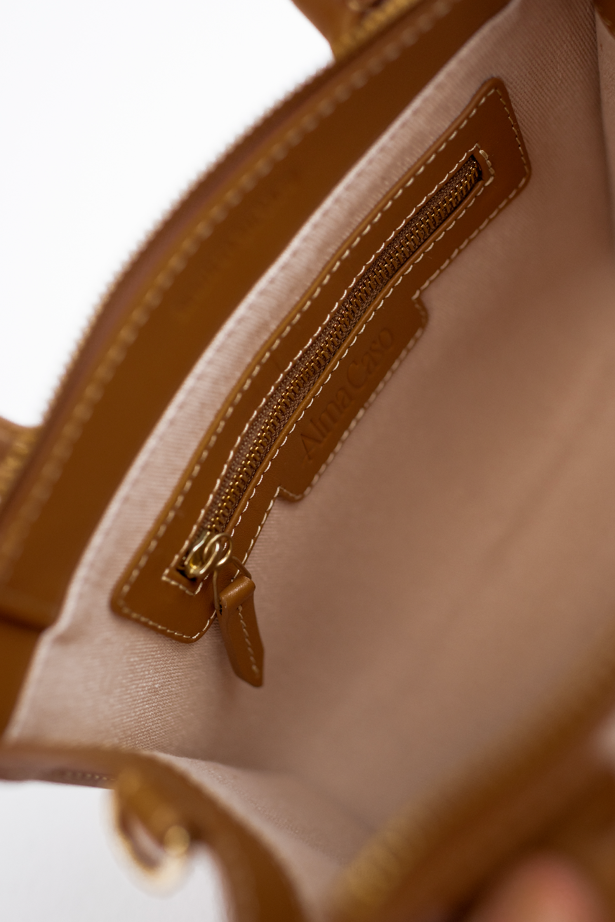 Close-up of the interior of a tan leather satchel bag showing a zippered pocket with gold-tone hardware and a beige fabric lining. The brand name 'Alma Caso' is embossed on the leather pocket.