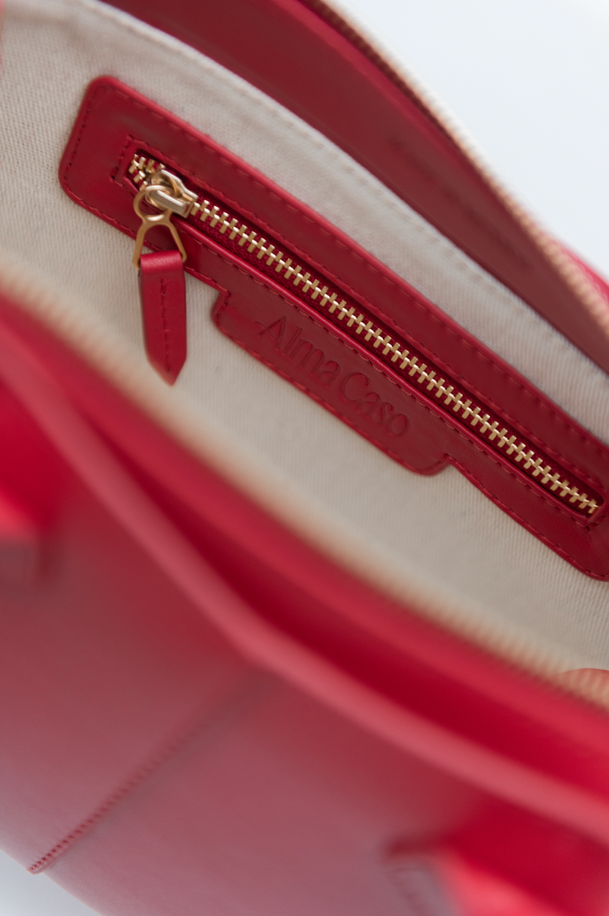 Close-up of the interior of a red handbag showing a red leather zipper pocket with a gold-tone zipper and the brand name 'Alma Caso' embossed on the leather.