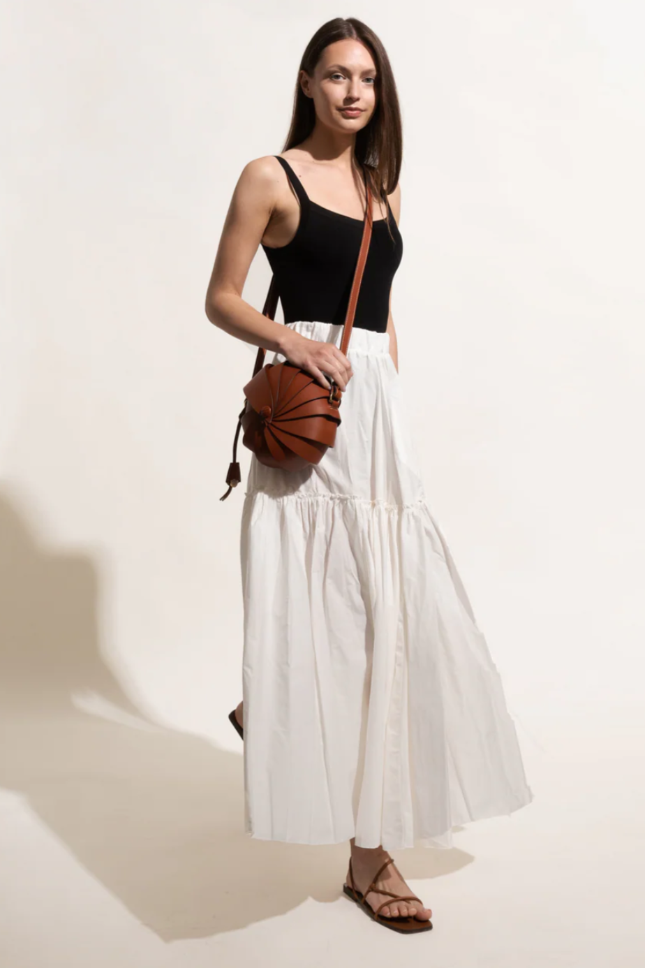 Woman wearing a black top and white skirt with leather sandals and a brown leather armadillo bag on a light background.