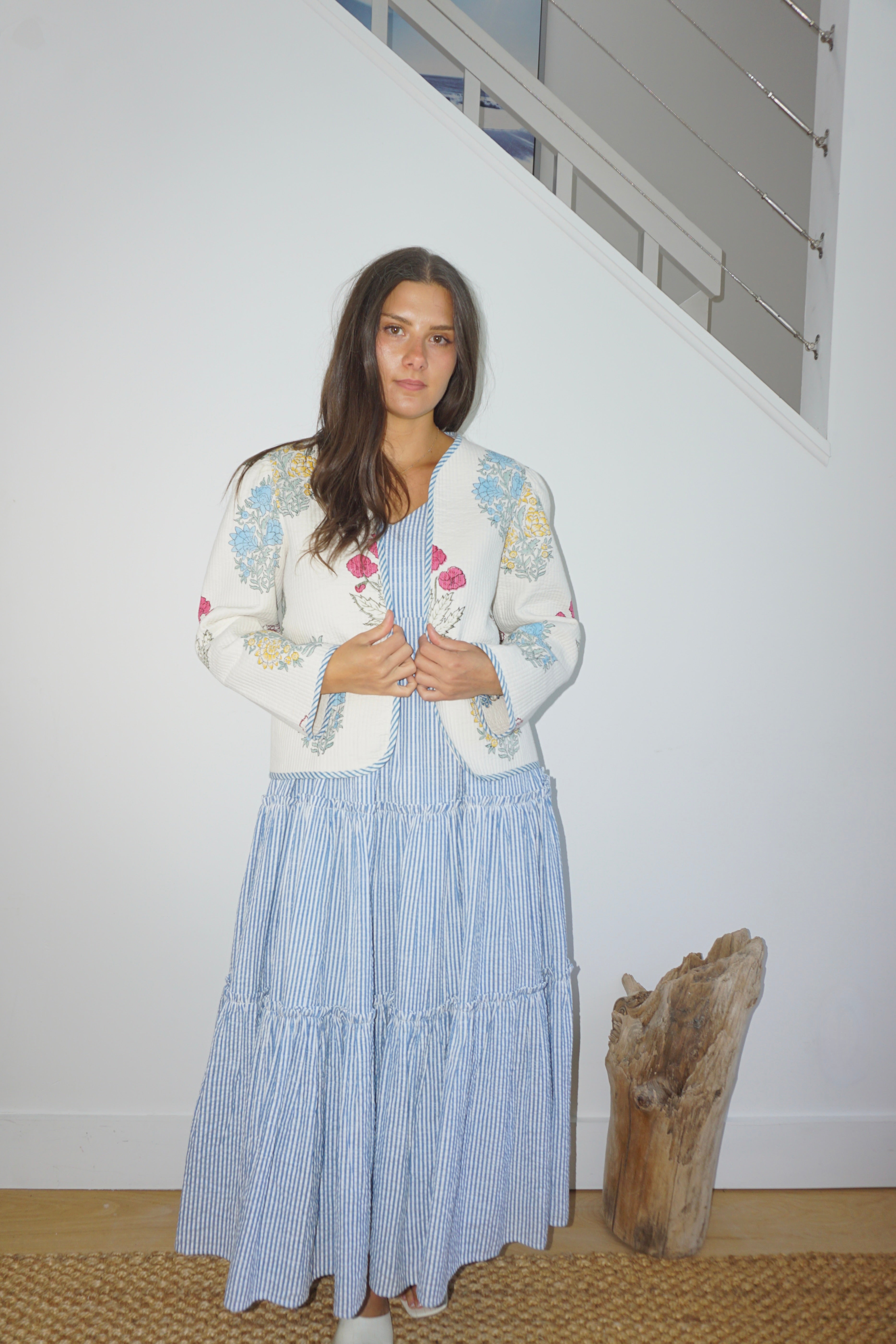 Woman wearing a white embroidered cardigan over a blue striped dress in a minimal indoor setting.