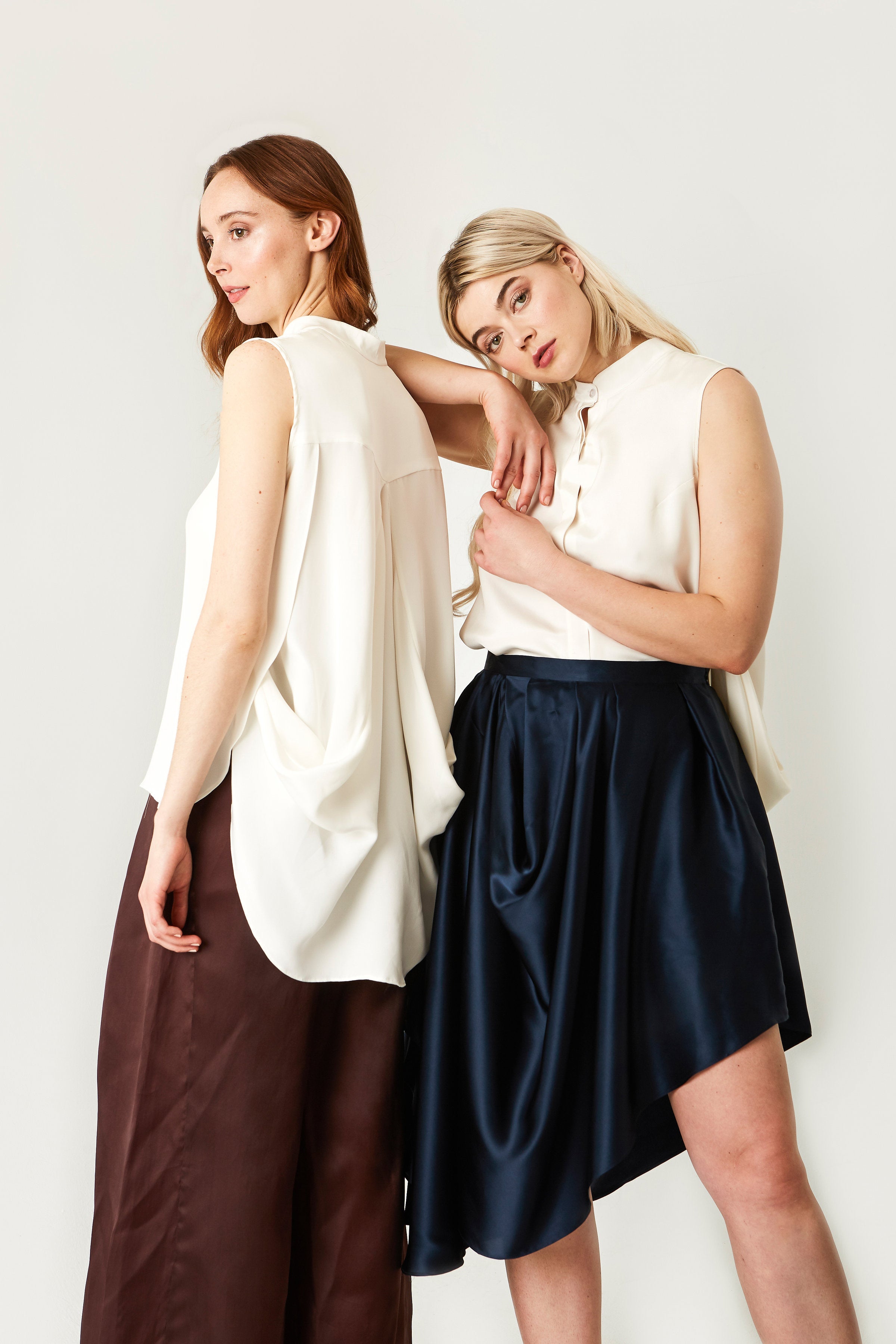 Two women wearing silk white sleeveless tops and dark pants and skirt against a plain background
