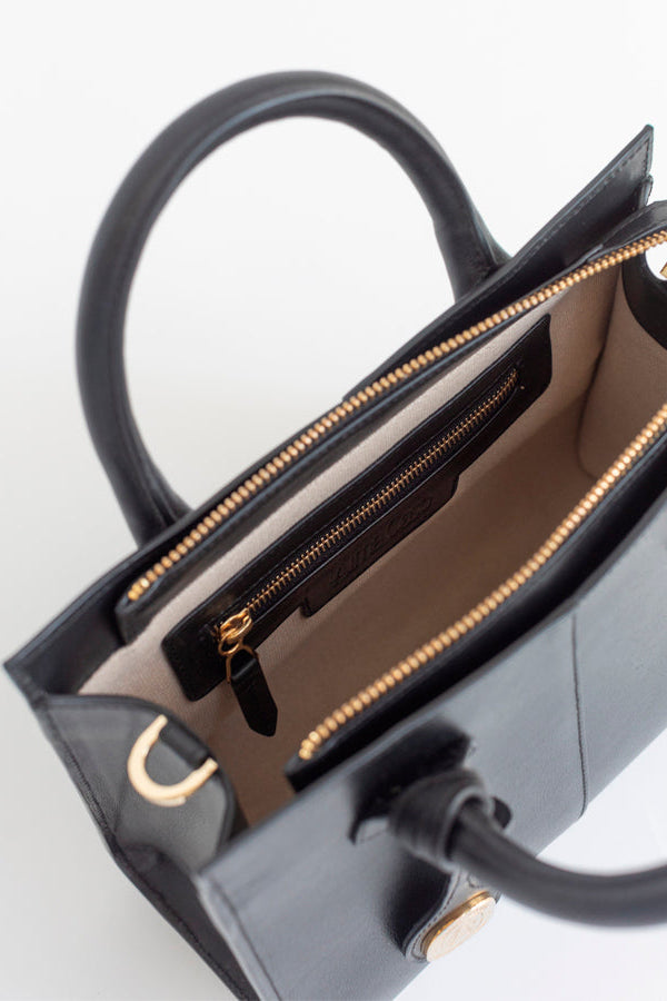 Close-up of a black leather handbag with gold-tone hardware, shown open to reveal a beige interior lining and zippered pocket.