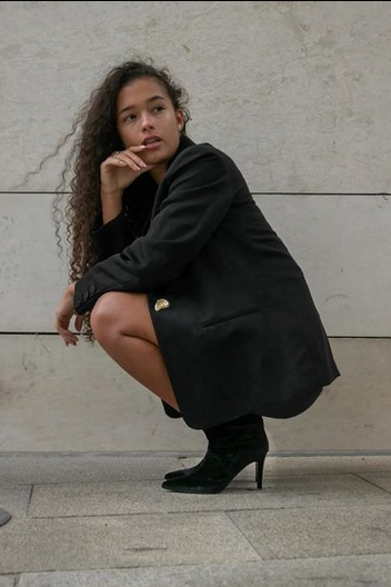Woman in a black coat and black shoes sitting against a light stone wall.