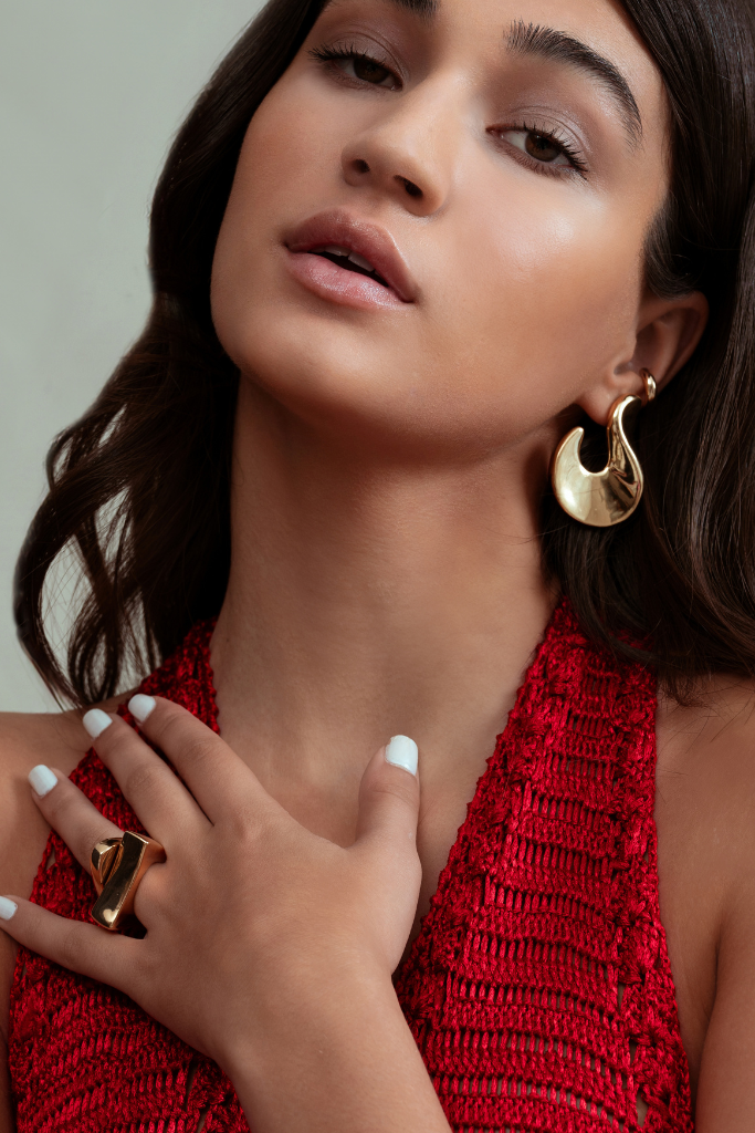 Woman wearing gold earrings and ring with a red top against a neutral background