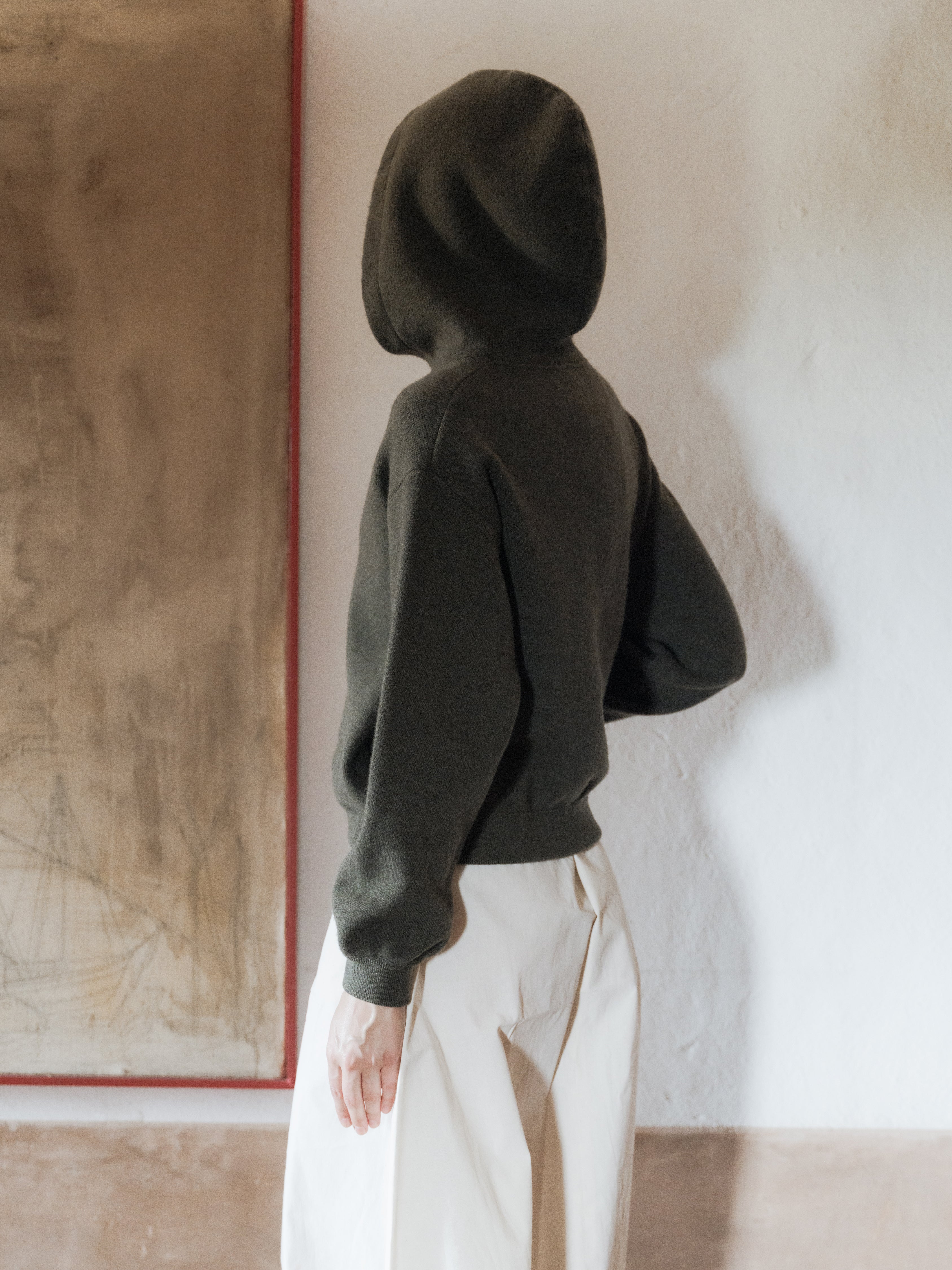 Person wearing a dark hoodie and white pants standing against a neutral wall.