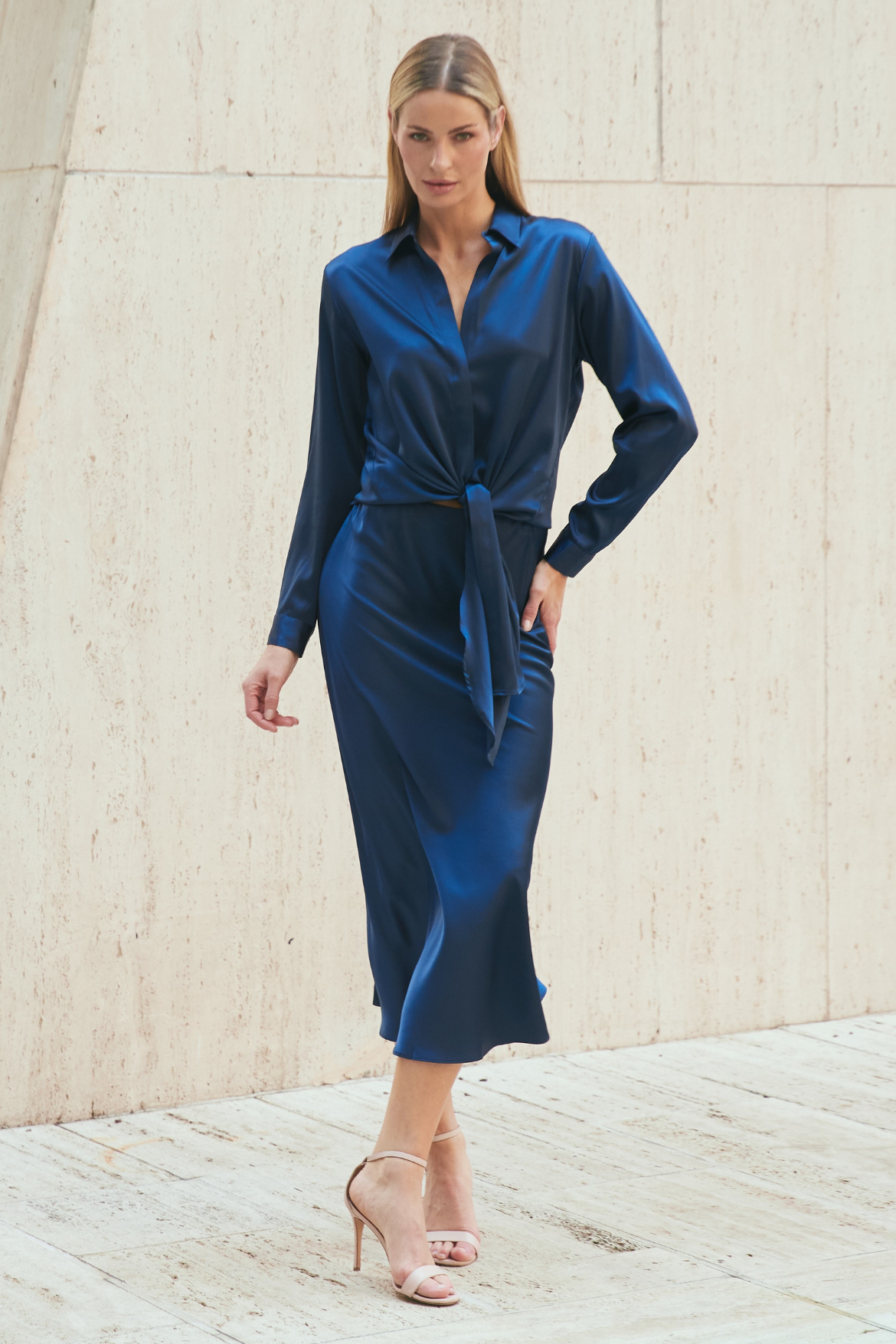 A woman modeling a dark blue satin silk midi shirt dress with a tie waist, standing against a light beige stone wall.