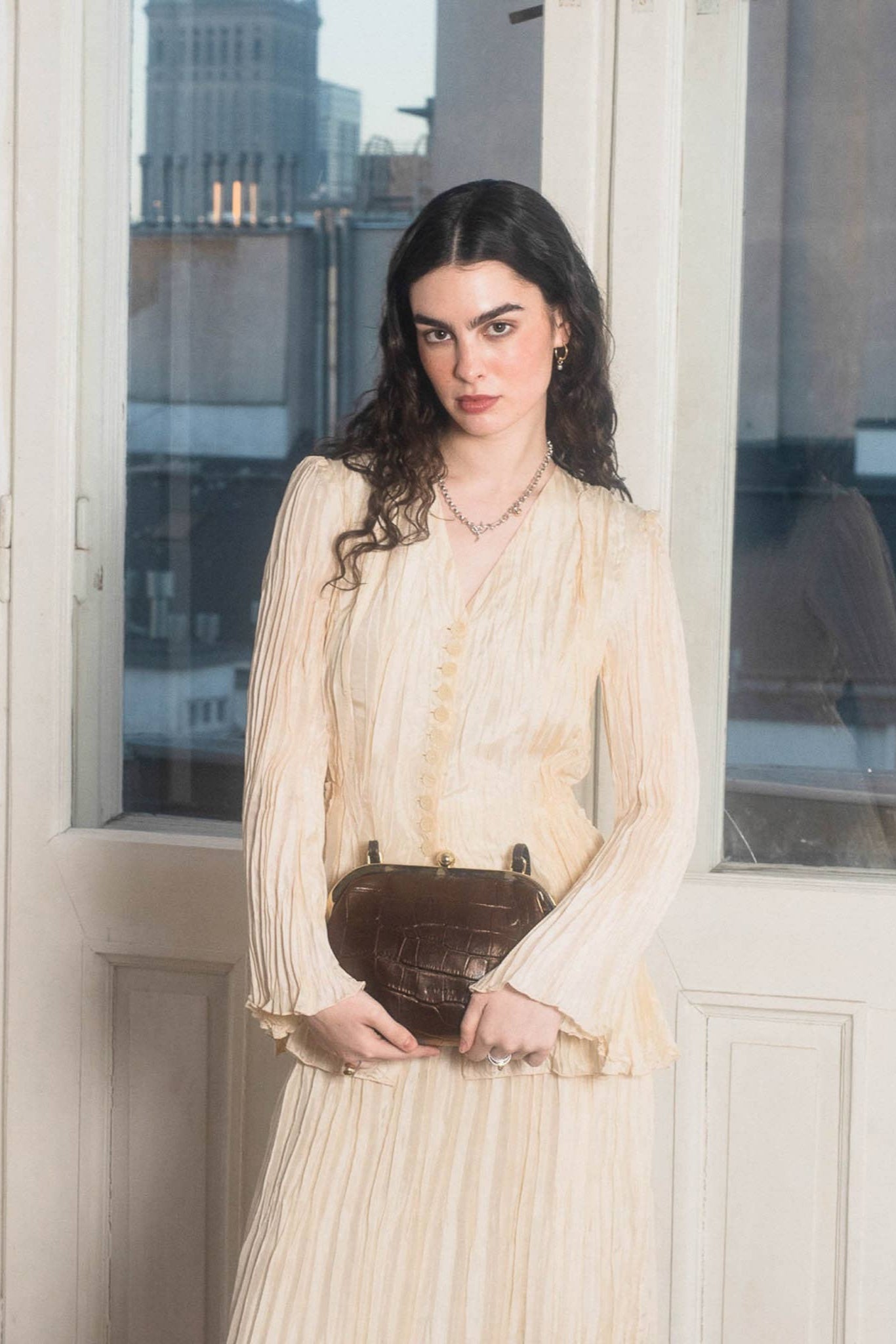 Woman in a beige dress holding a brown handbag indoors with a window view.