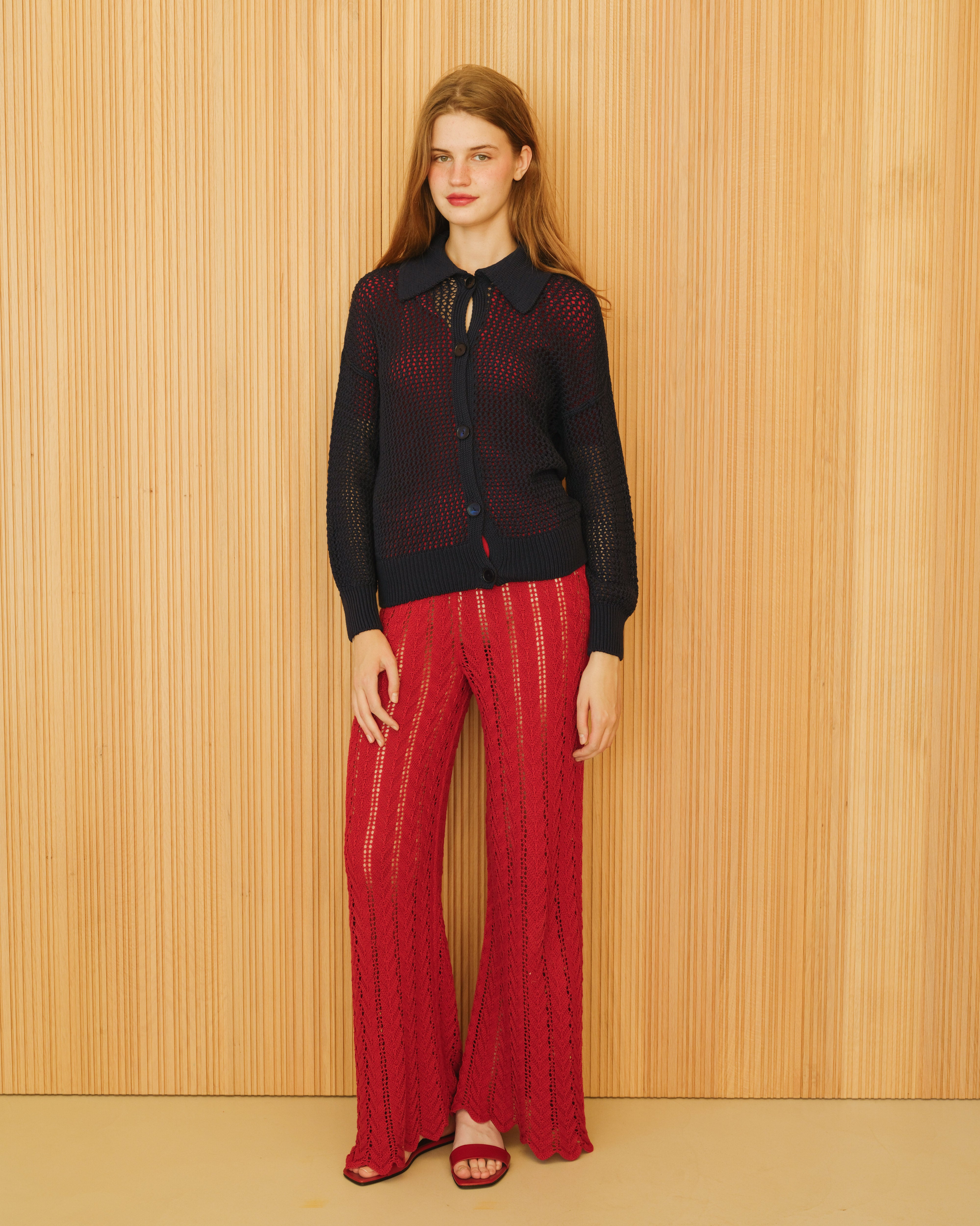 Woman wearing a black top and red pants against a wooden paneled wall.