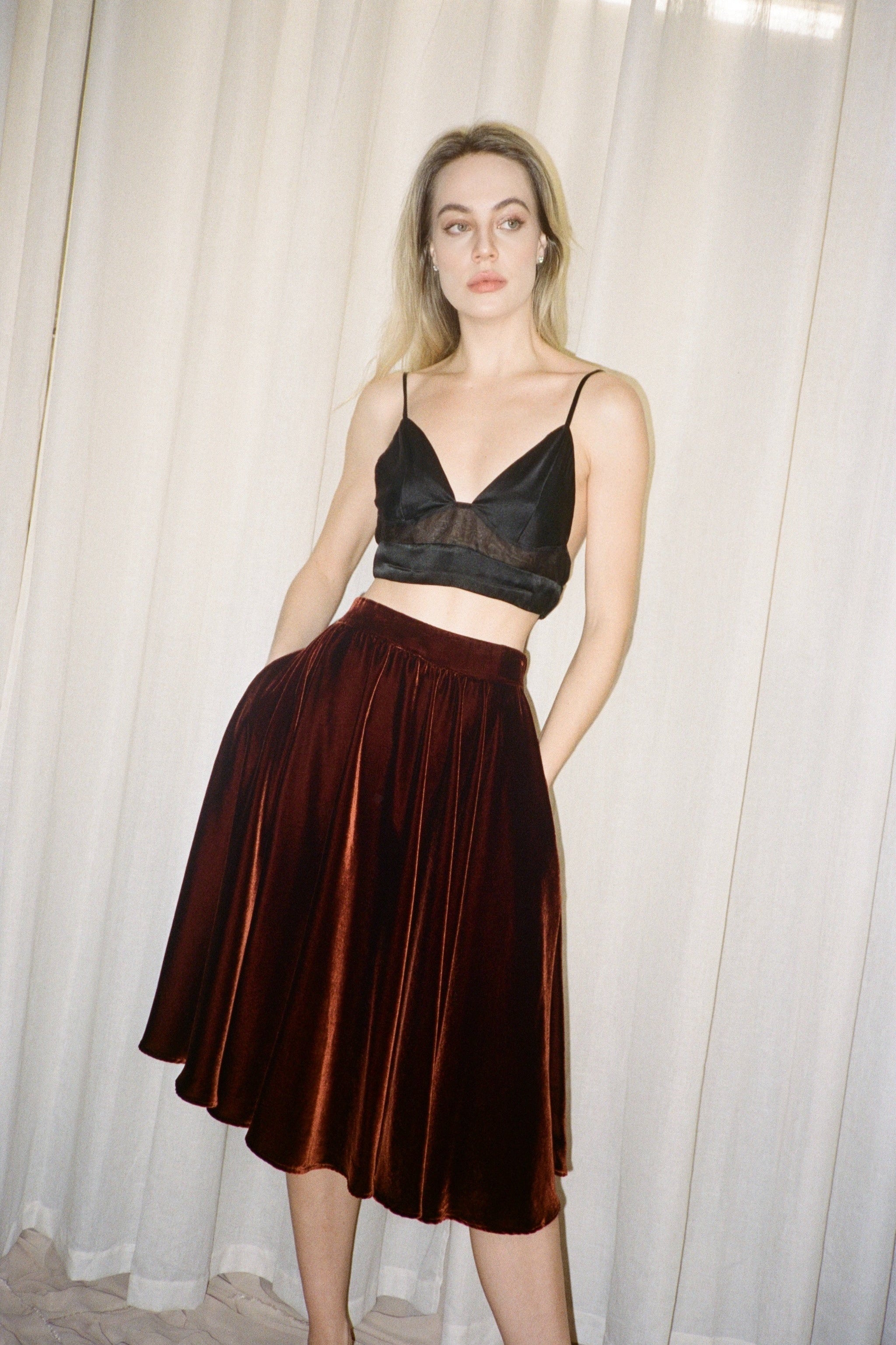 Woman wearing a black lace top and dark copper brown velvet skirt against a white curtain background