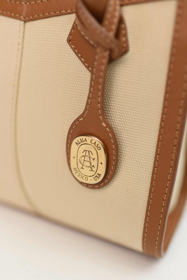Close-up of a beige canvas handbag with tan leather trim and a round gold-tone Alva Caso logo charm.