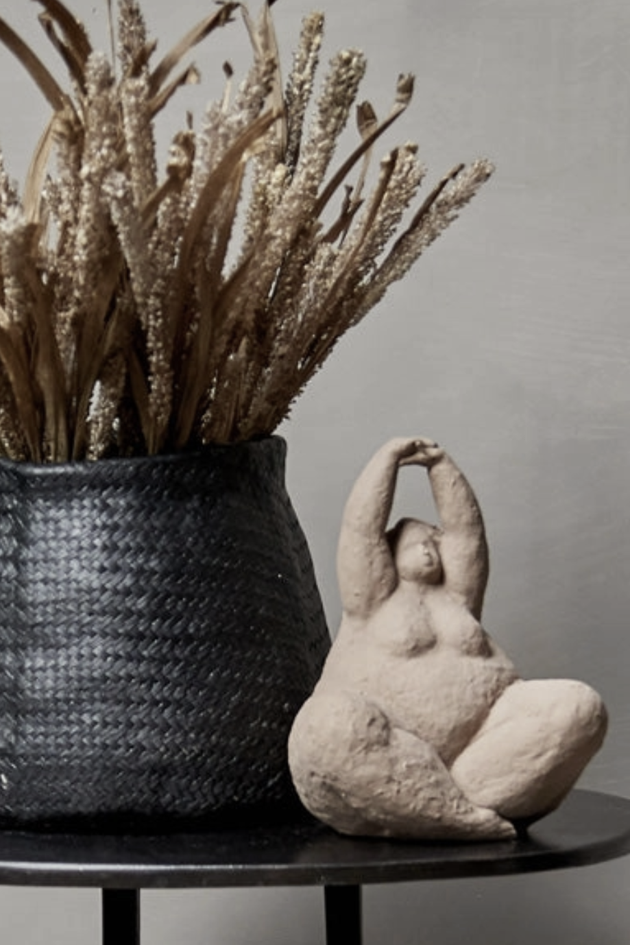 Decorative pot with dried plants and a stone sculpture of a woman on a table against a gray background