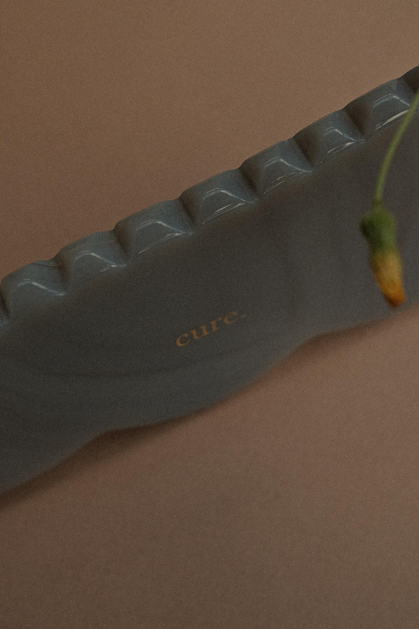 A gray silicone foot-shaped massage or reflexology tool with the brand name 'cure.' printed in gold on the side, resting on a solid brown background with a plant stem entering from the right.
