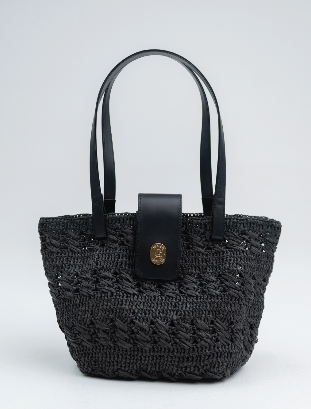 A black woven straw tote bag with black faux-leather handles and a central flap featuring a gold-tone crest logo, set against a light gray studio background.