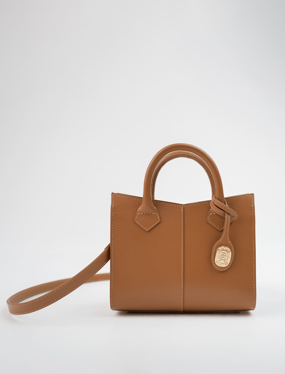 A structured tan leather handbag with dual top handles, a detachable shoulder strap, and a round hanging charm, photographed against a clean light gray studio background.
