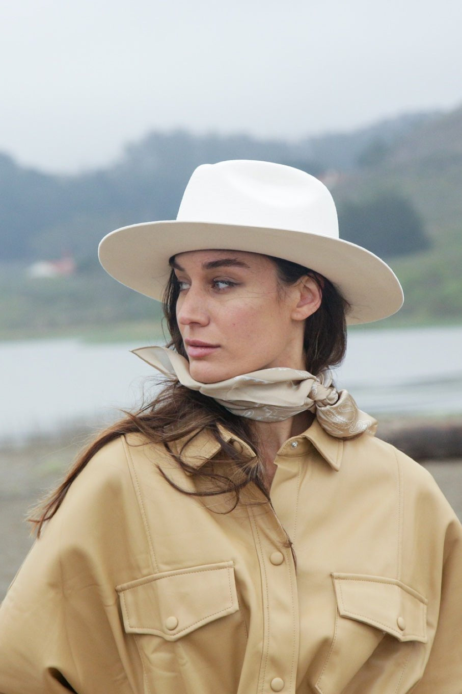 Woman wearing a beige jacket and white hat standing by a body of water with mountains in the background.