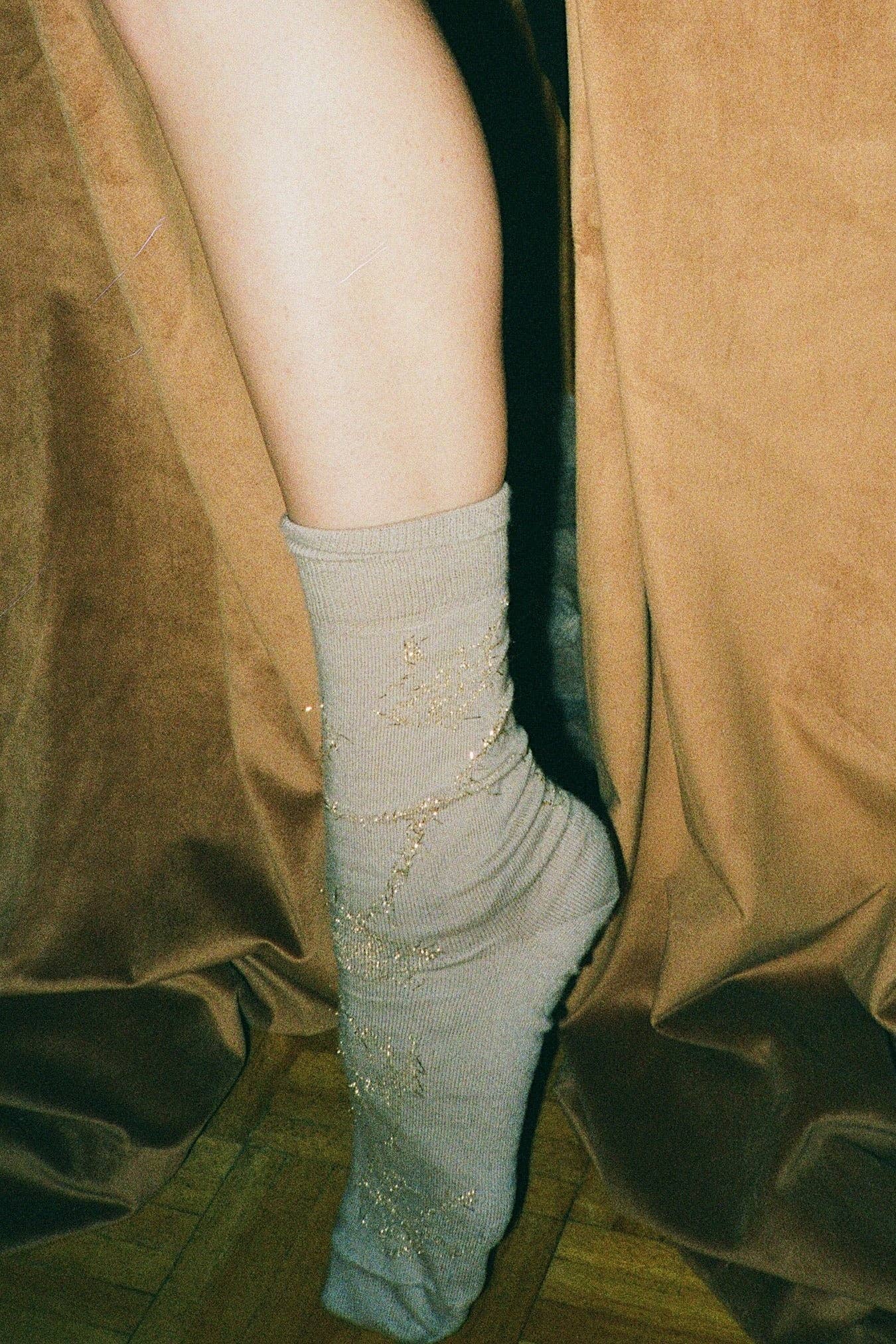 Leg wearing a light gray sock with decorative elements against a brown curtain background