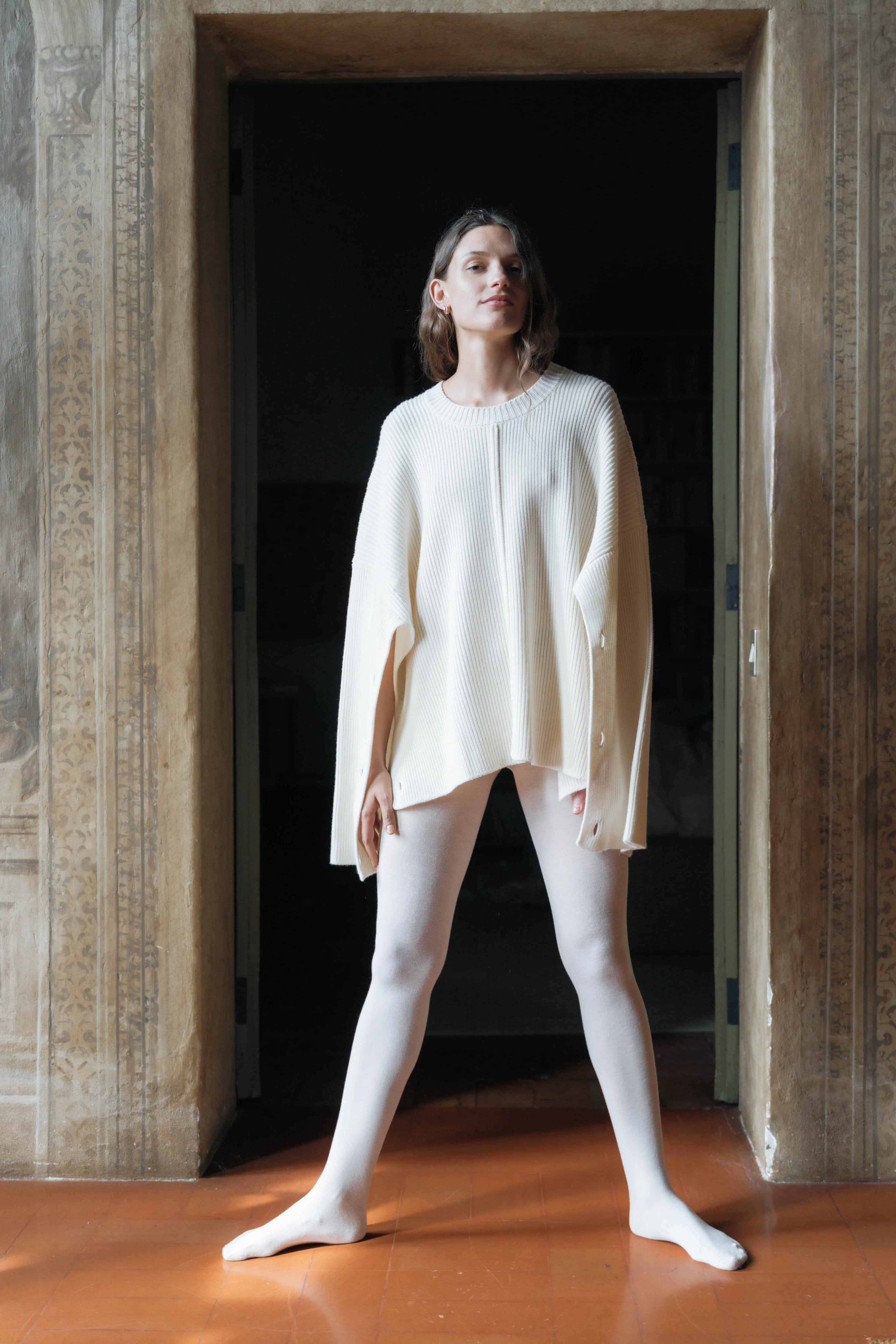 Woman wearing a cream-colored cape cardigan and tights standing in a doorway with a dark background