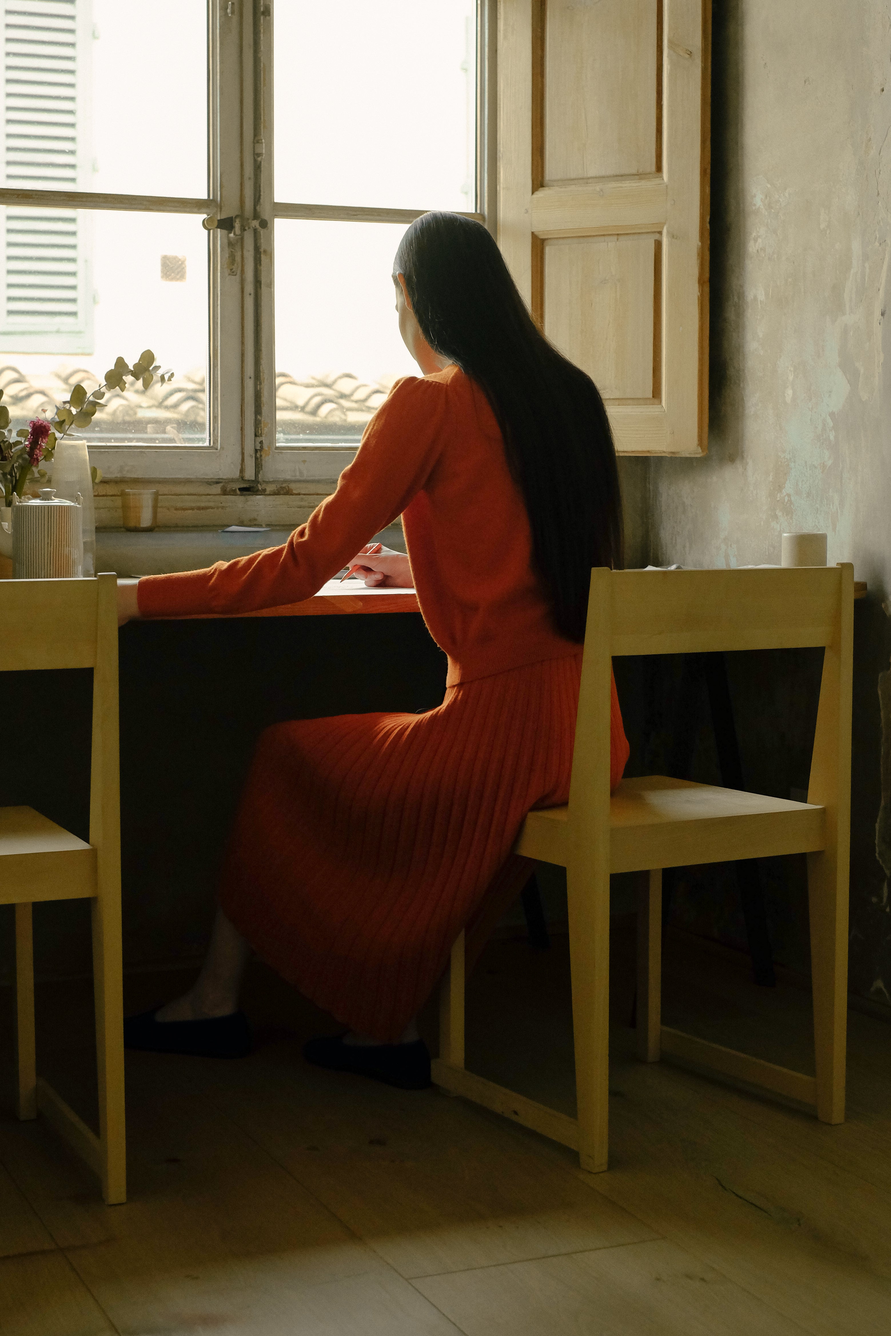 Woman with long brunette hair in an orange jumper and orange midi skirt sitting at a desk by a window in a dimly lit room.