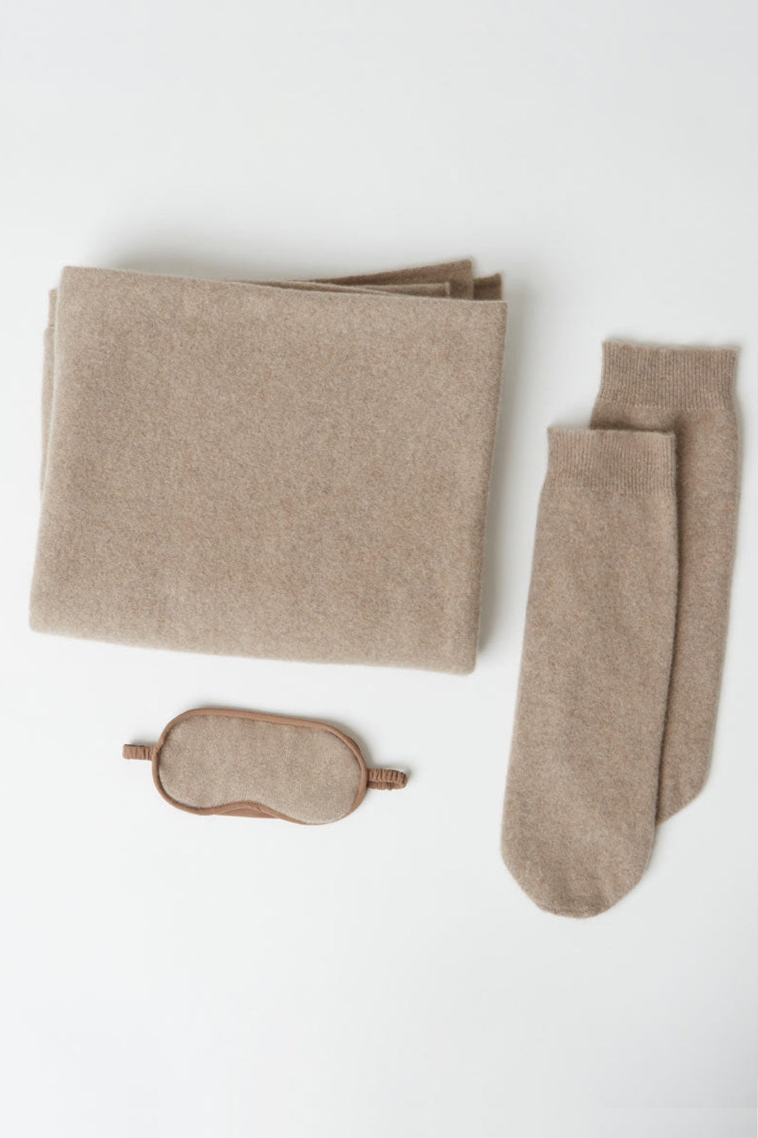 A cozy knit travel set in a warm taupe-beige color, including a folded blanket, a pair of socks, and a matching eye mask, arranged neatly on a white background.