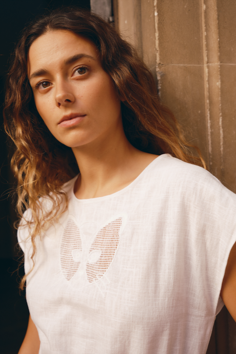 A woman wearing a white linen dress with a cat face embroidery on the chest, standing in front of a neutral background.