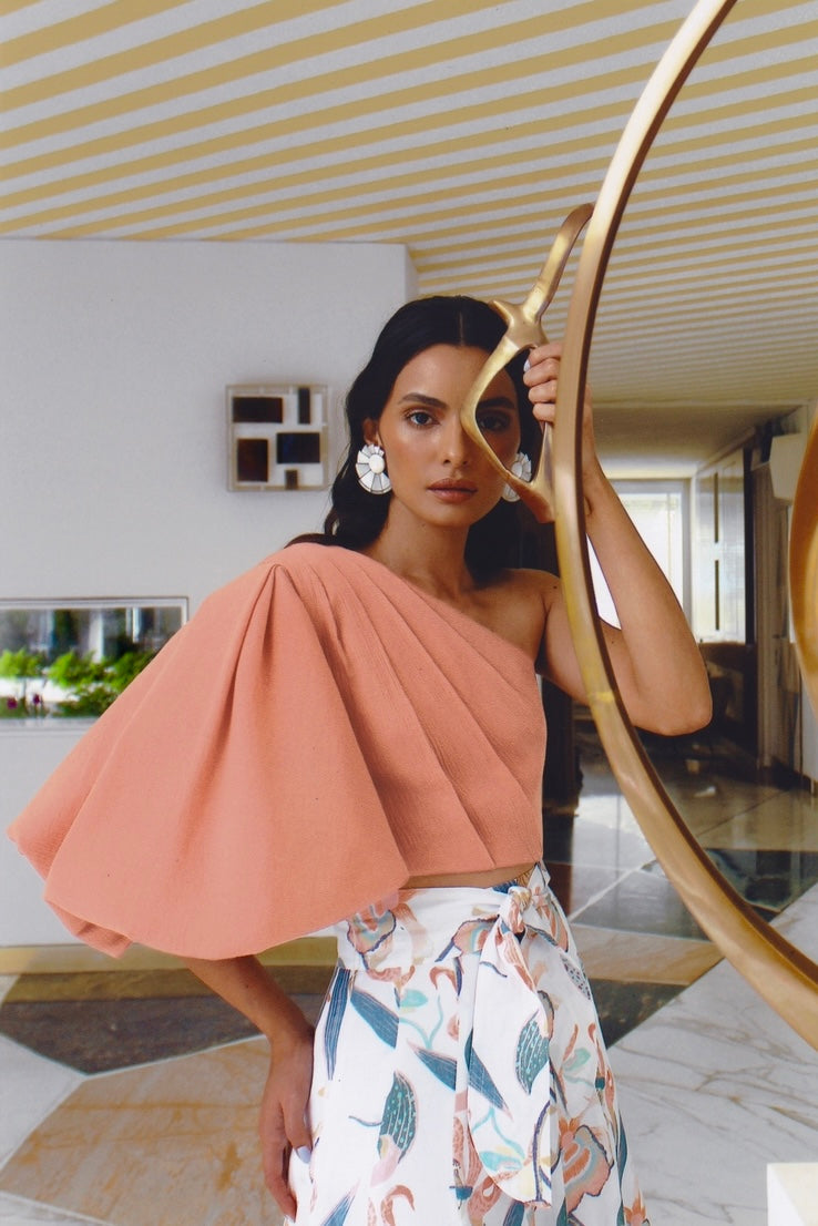 Woman wearing a peach coral colored one-sleeve top in a modern interior setting
