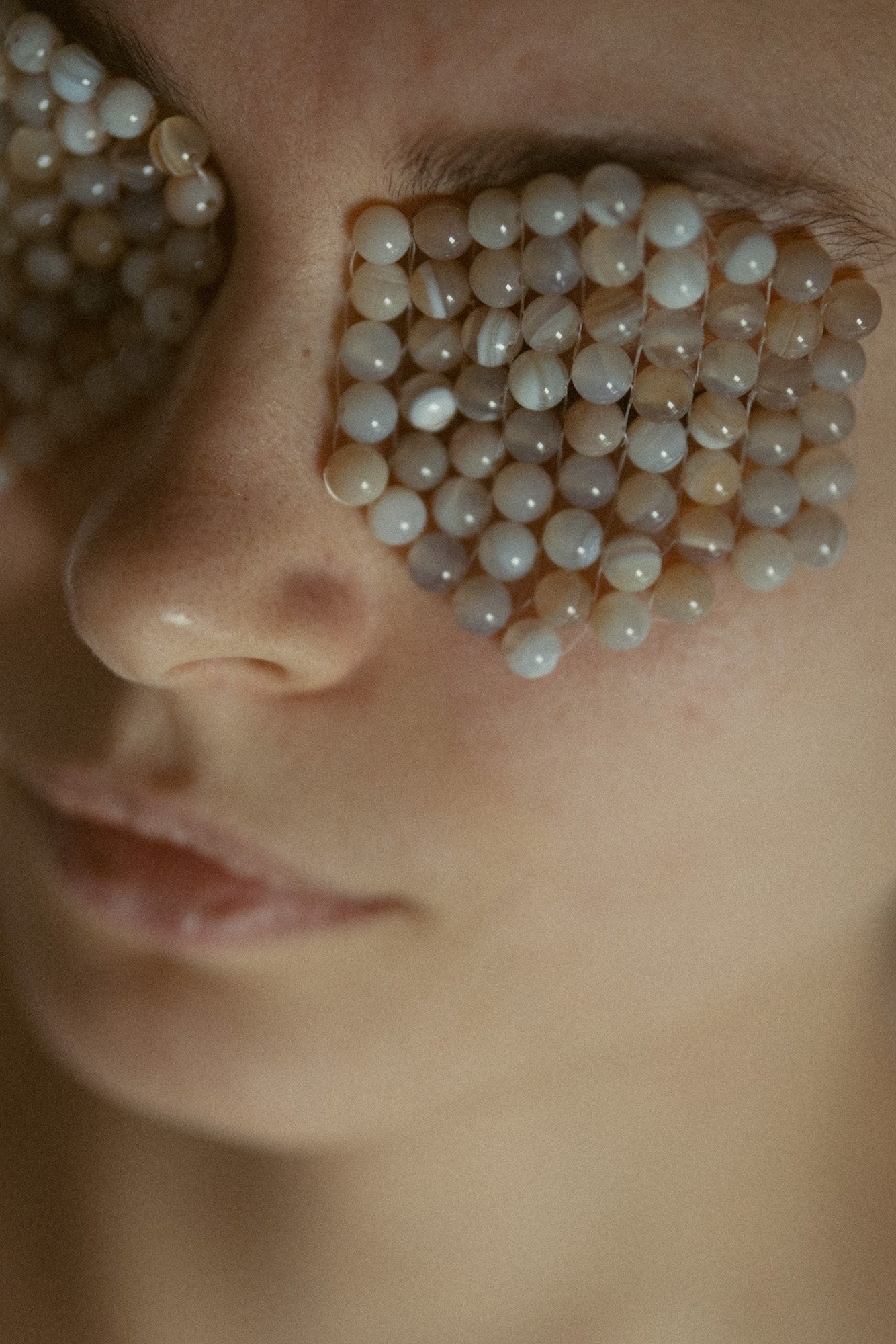 A close-up of a person's face with small white and beige pearl-like beads arranged across the eyes and cheek.