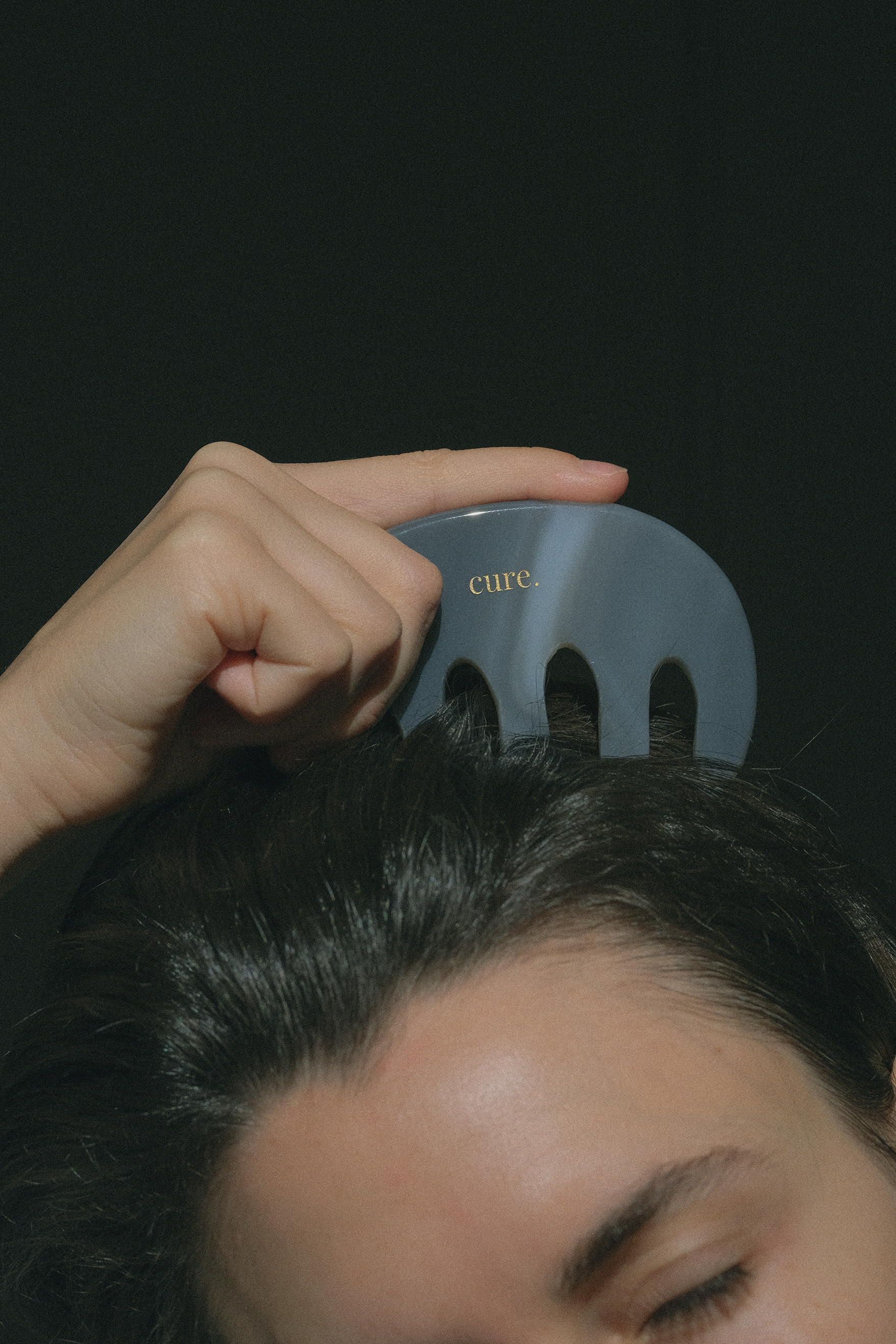 A close-up of a person using a gray plastic hair comb on their dark hair against a dark background.