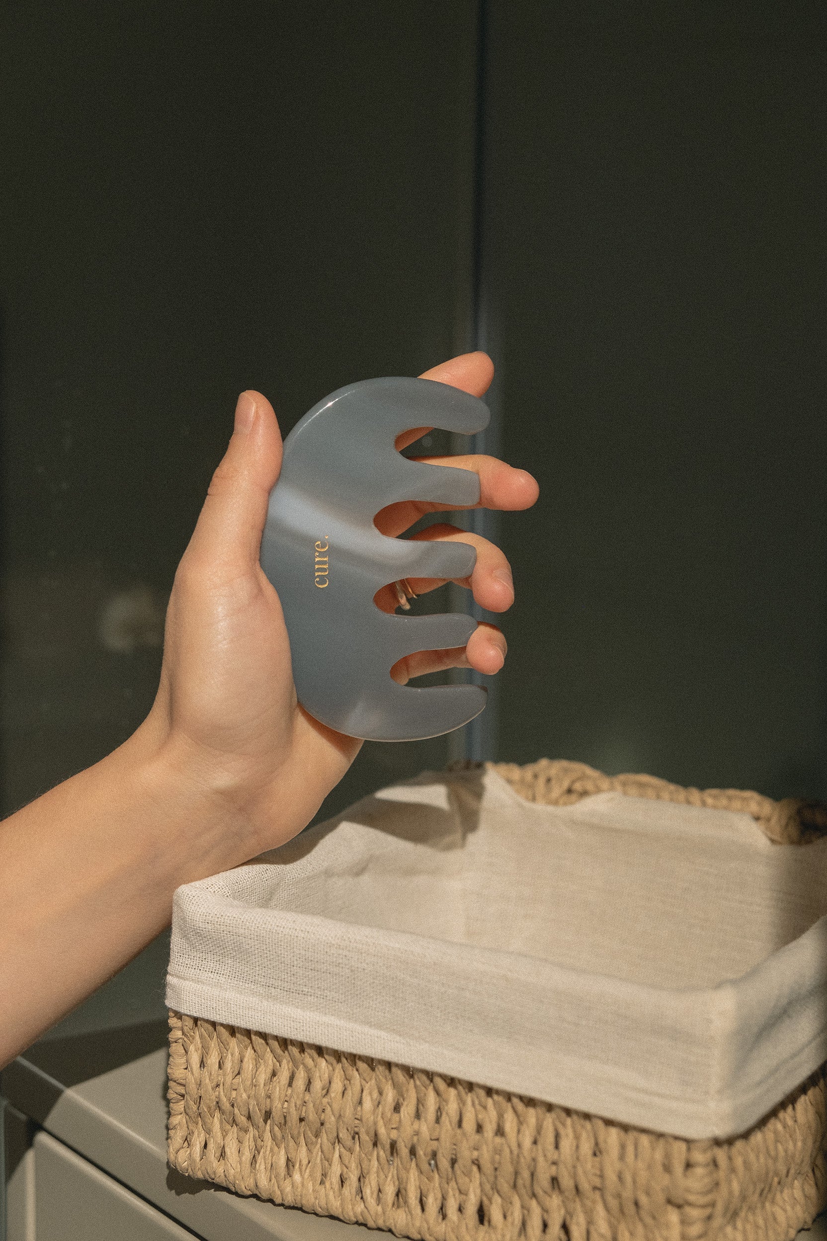 A hand holds a translucent gray hair claw comb over a woven basket lined with a white cloth, against a dark gray background.