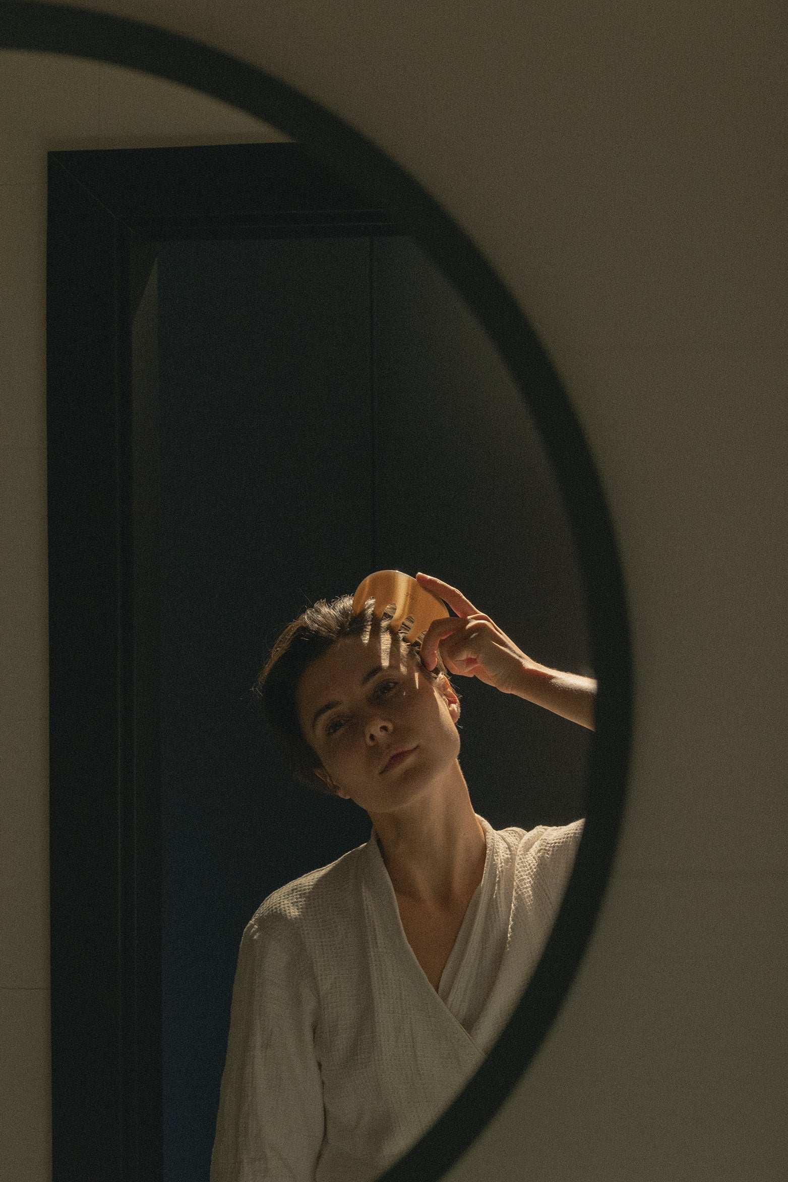 A woman in a white robe looks into a dark-framed oval mirror while holding a small gold object near her face in a dimly lit bathroom.