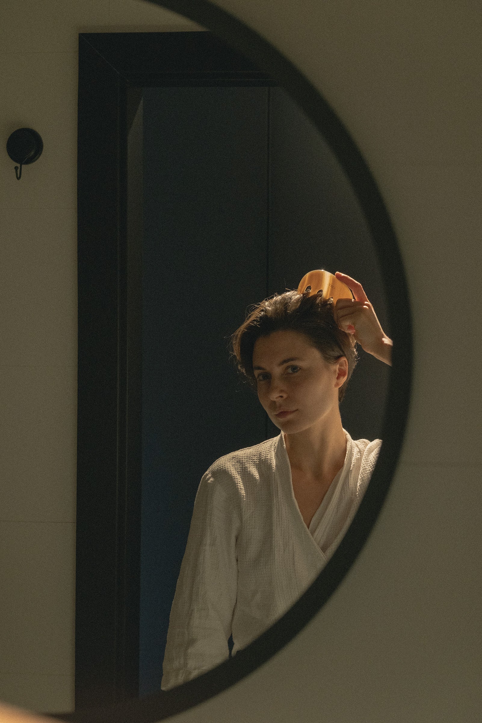 A woman in a white robe looks into a dark-framed oval mirror while adjusting her hair with a yellow hair accessory in a dimly lit room.