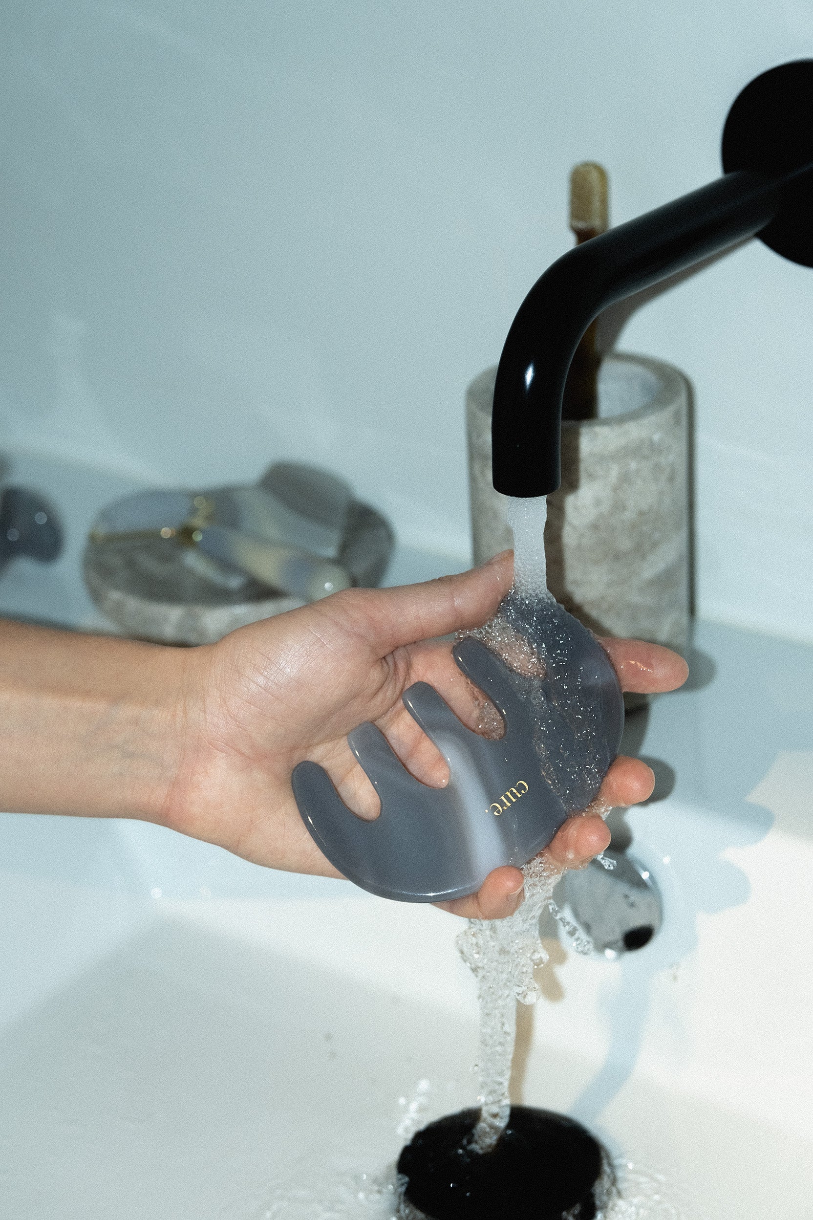 A hand holding a translucent gray silicone massage scraper under a running faucet in a sink, with a black faucet and a textured container in the background.