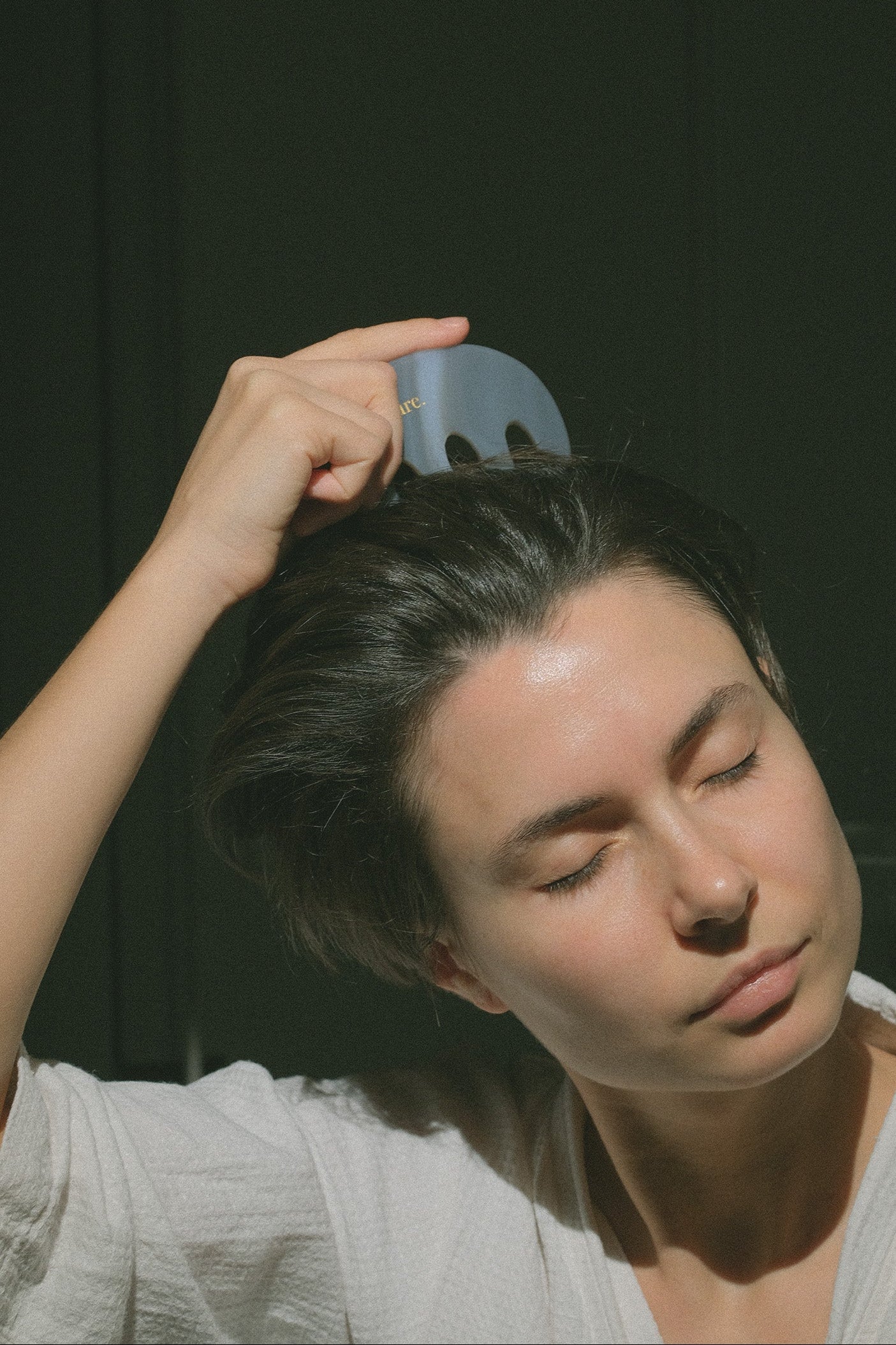 A person with short dark hair uses a light blue comb to detangle their hair, wearing a white textured top against a dark background.