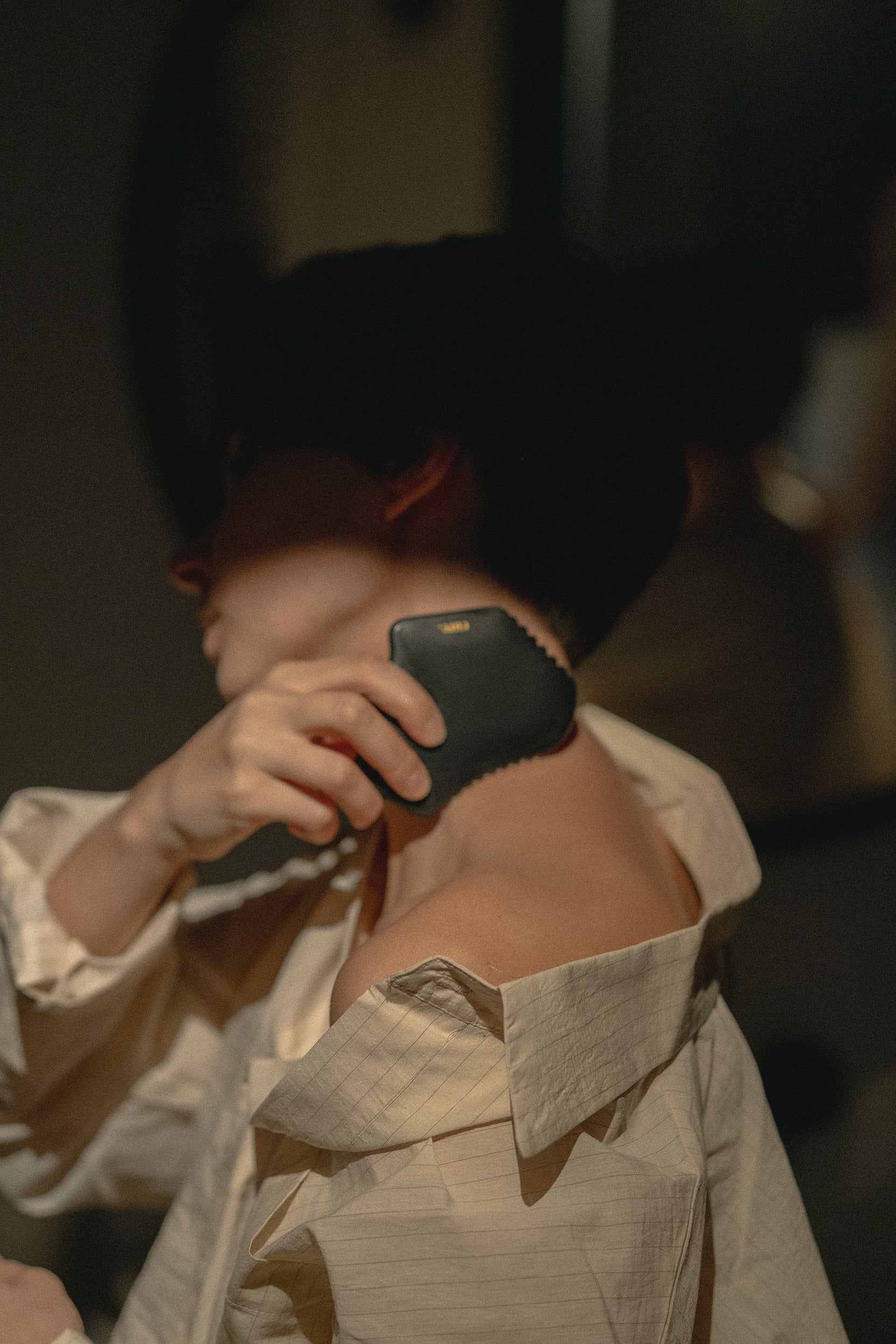 A person with dark hair wearing a light beige shirt uses a black agate body gua sha on their neck in a dimly lit indoor setting.
