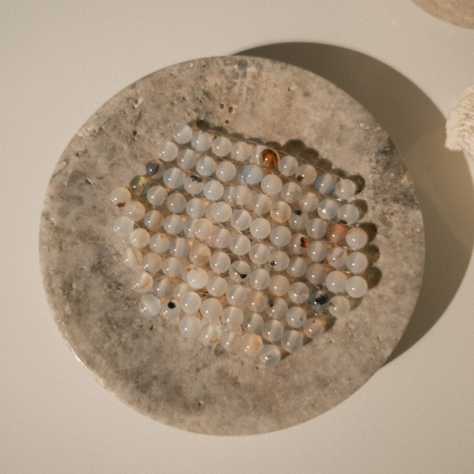 A top-down view of a stone eye patch made of small, round, milky white beads with subtle gray and brown speckles on top of a shallow circular dish on a light beige surface.
